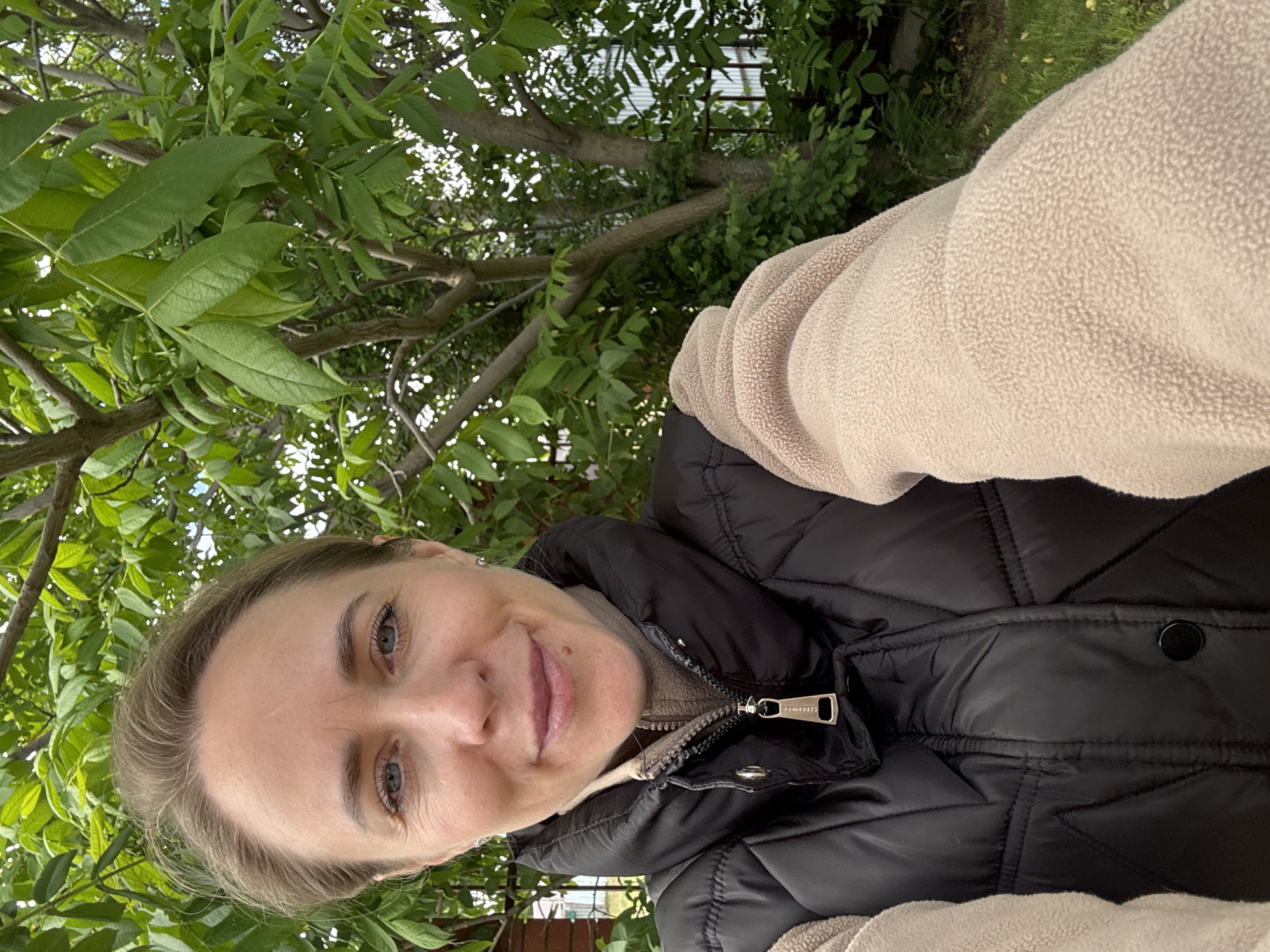 A woman in a black puffer jacket with a beige fleece underneath is taking a selfie outdoors. The background consists of green leaves and branches of trees. The location cannot be confirmed as Chaikovsky, Russia without additional information.