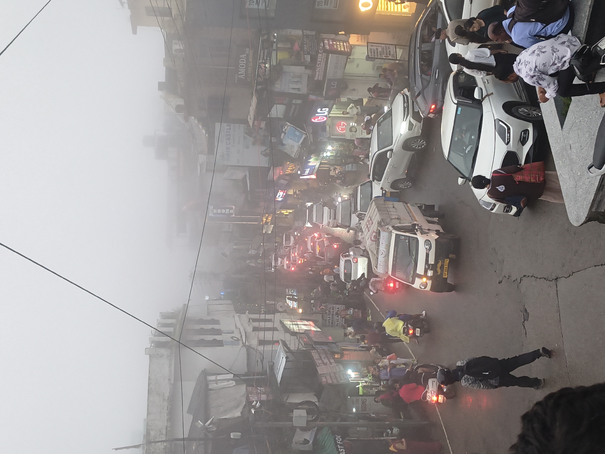 The image depicts a busy street scene in Dharamshala, India, characterized by heavy fog and traffic congestion. The narrow street is lined with shops and buildings, some displaying illuminated signs for businesses like "Hair Castle," "AMODA," and "LG." Vehicles, including cars, scooters, and a small truck, are seen in motion and stalled in traffic. Pedestrians are walking on the sidewalks and in the street, navigating the crowded conditions. The overall atmosphere suggests a typical day in a bustling urban environment, with the dense fog adding a slightly obscured and ethereal quality to the scene. The presence of various shops and the mix of local and modern vehicles indicate a commercial and residential area.