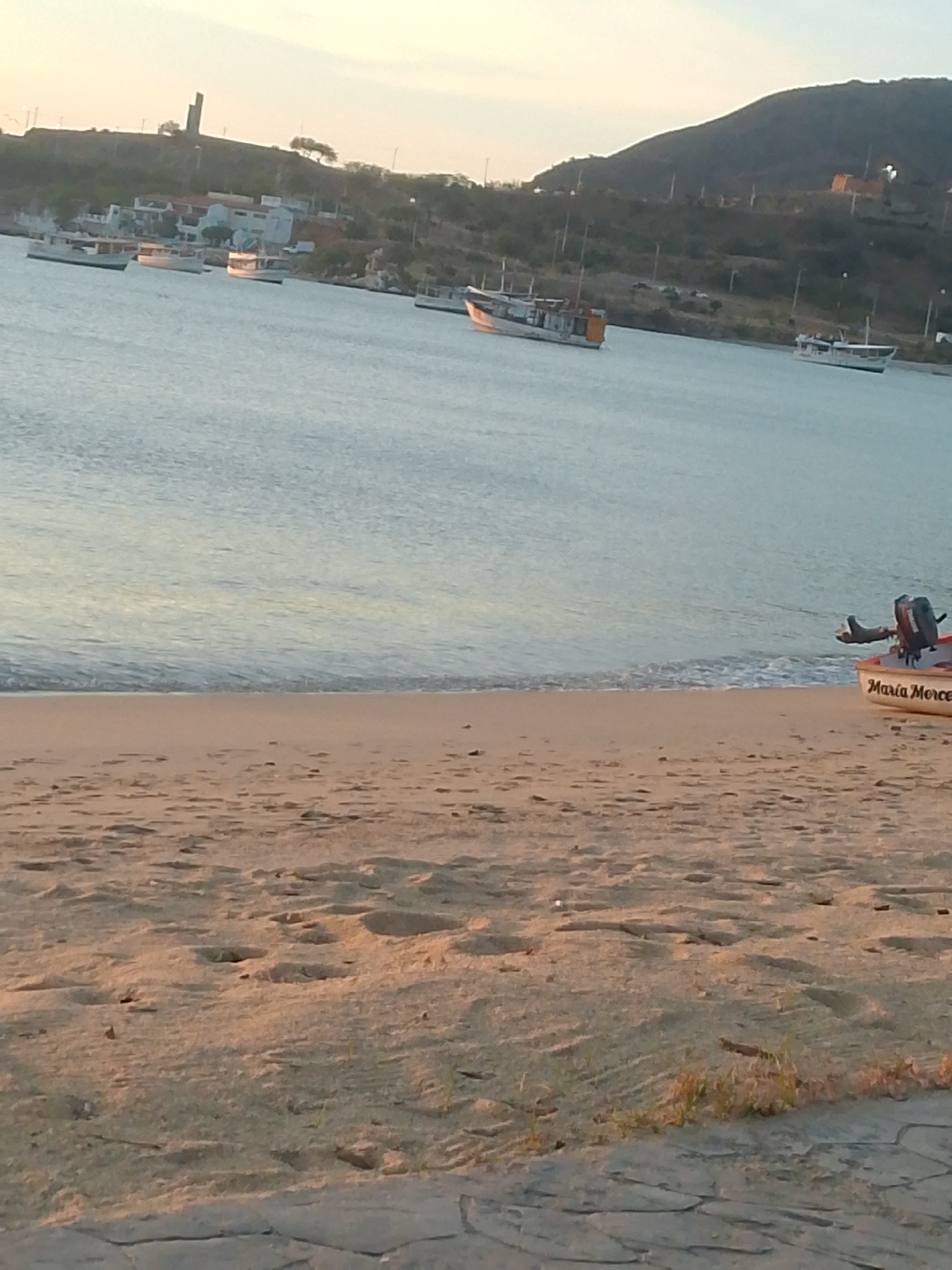 The image depicts a tranquil coastal scene in Juan Griego, Venezuela, likely captured during either sunrise or sunset given the soft, warm light. The foreground features a light brown sandy beach marked with numerous footprints and minimal sparse vegetation. To the immediate right, a dark, textured pavement section is visible, indicating the viewer's position.
In the midground, the calm, light blue-grey waters of a bay or inlet gently lap against the shore, with small ripples visible at the waterline. Partially beached on the far right, a small white boat named "María Merce" is visible, with its bow pointed left and an outboard motor attached to its stern, resting at the water's edge.
Across the bay, several small to medium-sized fishing or utility boats are anchored or floating. These vessels are predominantly light-colored, with one prominent boat displaying white and orange/brown sections.
The background consists of a hilly coastline. Buildings are visible along the distant shore and ascending the slopes, interspersed with green vegetation. A distinct tall, angular monument or structure, consistent with a fort or historical landmark, stands prominently on the leftmost hill. No individuals are visible in the scene.