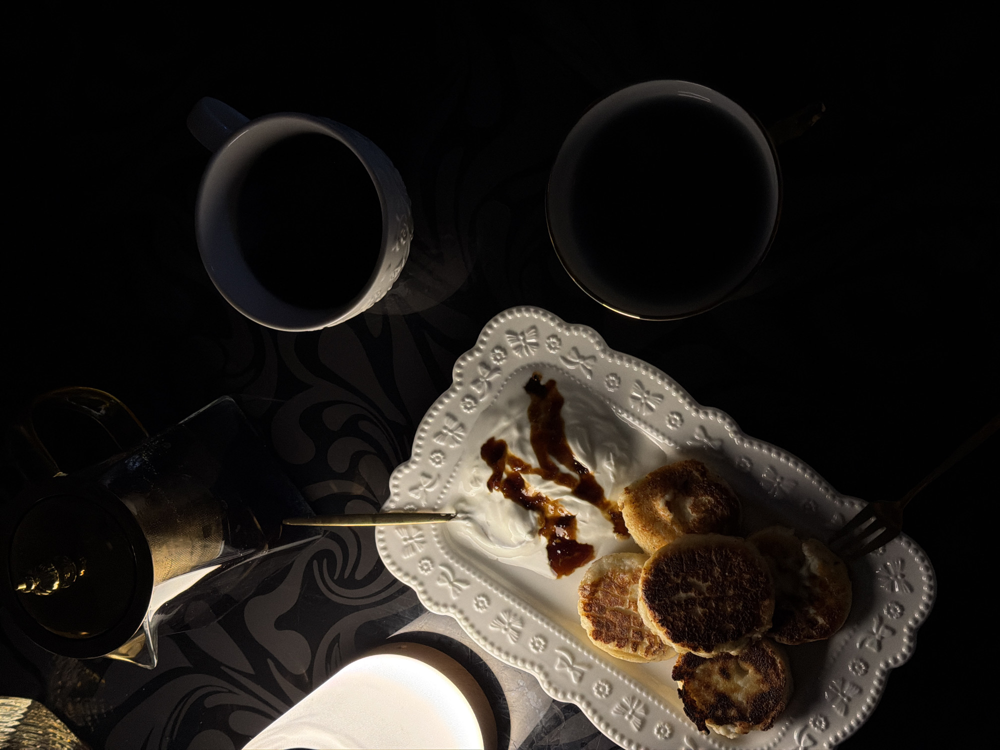 The scene is a low-lit, high-angle shot of a table setting. Two cups containing a dark liquid (likely coffee) are positioned at the top of the frame. A white, ornate plate sits below the cups, holding several small, round, cooked items resembling pancakes or fritters, topped with a dollop of white cream and streaks of a brown liquid. A gold-colored fork rests on the edge of the plate. To the left, there's a coffee pot, also gold-colored, sitting on a patterned surface. A lamp with a warm glow is positioned near the bottom of the frame. The location is Rossiyskiy, Russia.