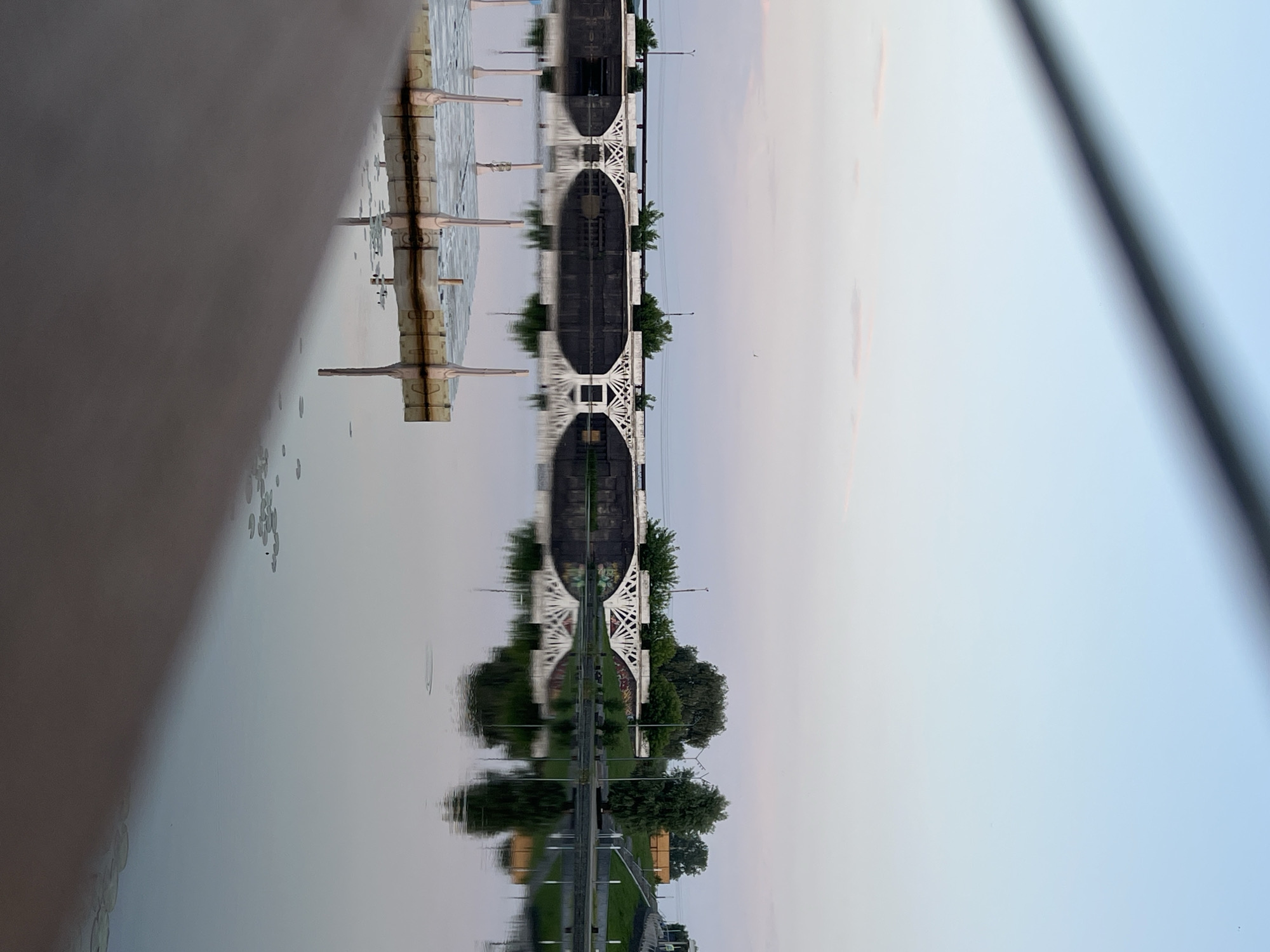 A horizontal bridge with decorative white arches is reflected in a still body of water. A floating dock with vertical posts is partially visible in the foreground, on the left side of the image. The sky above the bridge transitions from a pale pinkish hue near the horizon to a light blue at the top, with wispy clouds. Trees and some buildings are visible on the far bank of the water, behind the bridge. The scene appears to be captured at dawn or dusk, given the soft lighting and pastel colors of the sky. The location is Votkinsk, Russia.