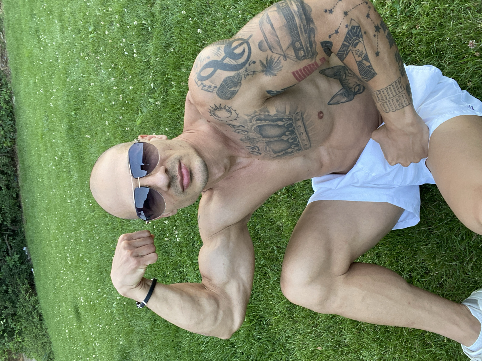 A bald, shirtless man with sunglasses is sitting on a grassy lawn. He is flexing his bicep and has tattoos on his chest and arm. The man is wearing white shorts and white sneakers. The location is an outdoor grassy area, possibly a park or backyard, with lush green grass and some small white flowers scattered throughout. The background shows more greenery, suggesting a natural outdoor setting. There is no indication of Pleven, Bulgaria in the image.