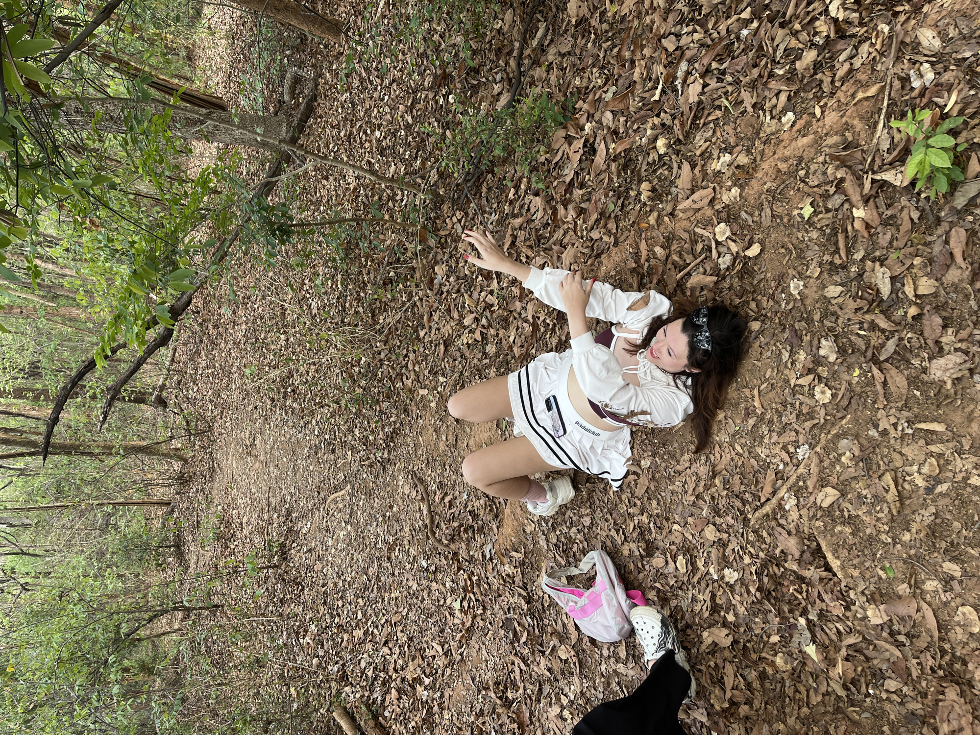 A woman is lying on a path in a wooded area in Chiang Mai City Municipality, Thailand. The ground is covered in fallen leaves. The woman is wearing a white outfit consisting of a short skirt and a long-sleeved top, and sunglasses are placed on her head. A white and pink bag is near her feet. There is another person wearing black pants standing near the woman. The location appears to be a walking trail in a forested area.