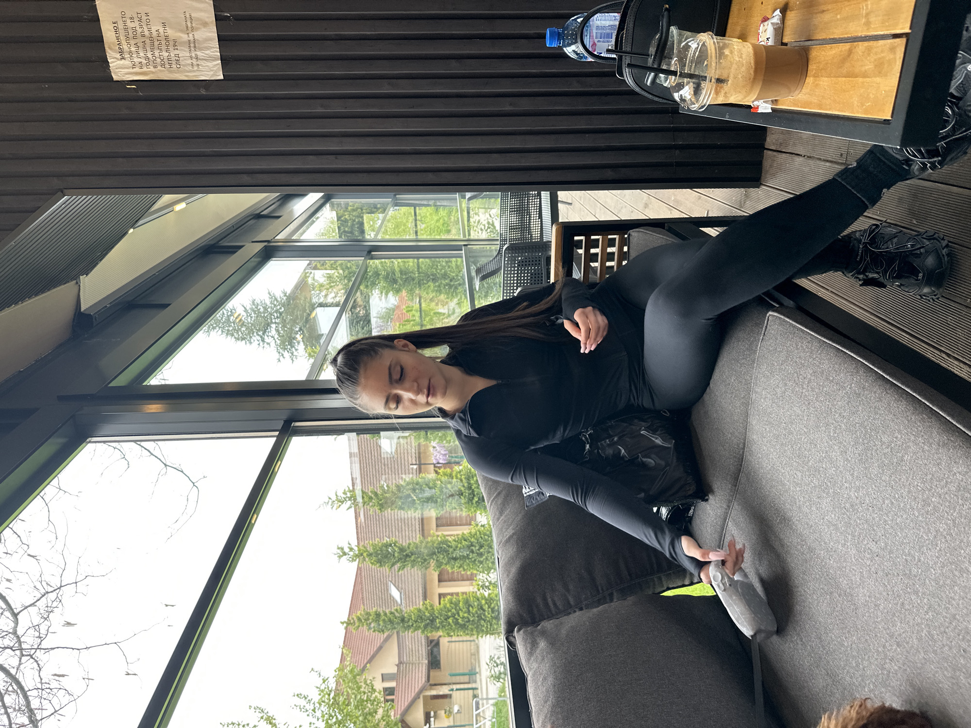 A young woman with long dark hair, pulled back in a ponytail, is seen relaxing on a grey couch in what appears to be a modern indoor-outdoor space, possibly a cafe or sunroom. She is dressed in a black long-sleeved top and black leggings, and her feet, clad in black Balenciaga sneakers, rest on the couch. Her right hand holds a grey remote control-like device. Her eyes are closed or looking down, suggesting a moment of rest or contemplation.
The setting features large glass windows that showcase a view of lush green trees and residential houses outside, hinting at a suburban or green urban environment. The interior has dark wooden slatted walls or ceiling, and a wooden table beside the couch holds a plastic water bottle with a blue cap, a partly consumed iced coffee in a clear plastic cup, and a black handbag.
The weather outside appears to be overcast or bright, without direct sunlight. On the wooden slatted wall above, a white paper sign with Bulgarian text is visible. The OCR translation of the sign reads: "SMOKING IS PROHIBITED FOR PERSONS UNDER 18 YEARS OF AGE IN THE PREMISES AND ACCESS FOR MINORS AFTER 19:4! IN CASE OF NON-COMPLIANCE WITH THE PROHIBITION, THE POLICE WILL BE CALLED!" The mention of "19:4" might be a typo in the original sign or OCR for "19:00" or similar.
The overall scene suggests a relaxed atmosphere in an airy, contemporary space, possibly in Plovdiv, Bulgaria, given the location context.