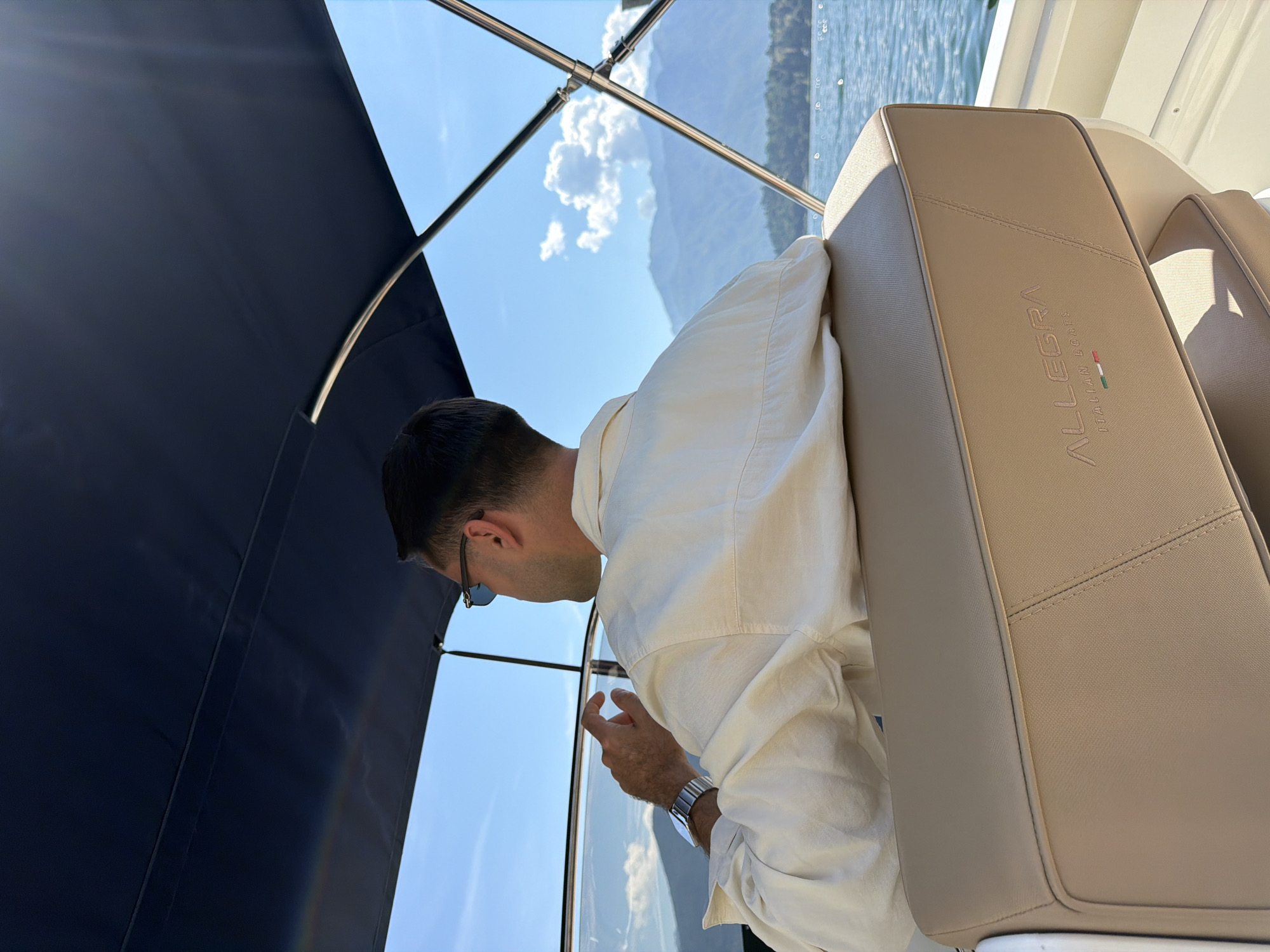 A person wearing a light-colored shirt and sunglasses is seated in the captain's chair of a boat, looking out at a body of water with mountains in the background. The boat appears to be on Lake Como, specifically near Tremezzina, Italy, given the "ALLEGRA ITALIAN BOATS" branding visible on the chair. The sky is blue with scattered clouds, and the sun is casting light on the scene. The person's left hand is visible, resting on the edge of the boat's console, with a wristwatch on their left wrist. The water is calm with small ripples.