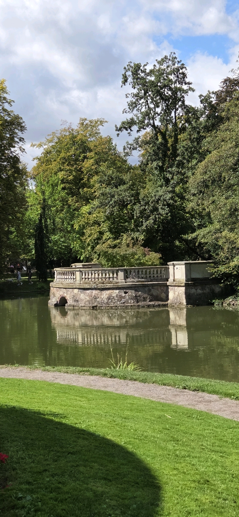 This image captures a serene park scene with a pond and a stone structure on the water. The pond is a murky green, reflecting the trees and the sky. A circular stone structure, resembling a small island, sits in the middle of the pond, topped with a decorative stone balustrade. The structure has arched openings at its base, suggesting it might be a fountain or a historical relic. Lush green grass surrounds the pond, with a gravel path winding through it. In the background, a dense forest of mature trees with vibrant green foliage provides a natural backdrop. The sky is partly cloudy with patches of blue. A few indistinct figures can be seen in the distance, walking on a path near the trees, implying the location is a public park. The overall atmosphere is tranquil and picturesque. This scene is located in the Parc de l'Orangerie in Strasbourg, France.