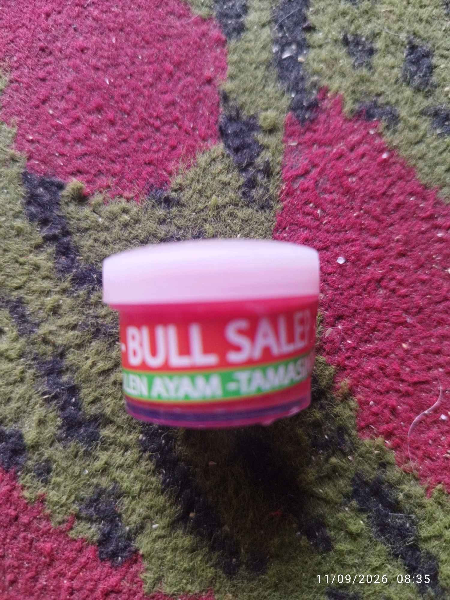 A small, pink-lidded container with a red label is placed on a textured surface that appears to be a rug with a pattern of dark red, green, and black patches. The label on the container is mostly obscured but displays white text that reads "BULL SALE" and "LEN AYAM-TAMAS". The container is positioned slightly off-center to the right. In the bottom right corner of the image, a timestamp "11/09/2026 08:35" is visible. The overall scene is poorly lit and out of focus, suggesting a casual, non-professional photograph. No specific location in Mojokerto, Indonesia, can be determined from the visual information provided.