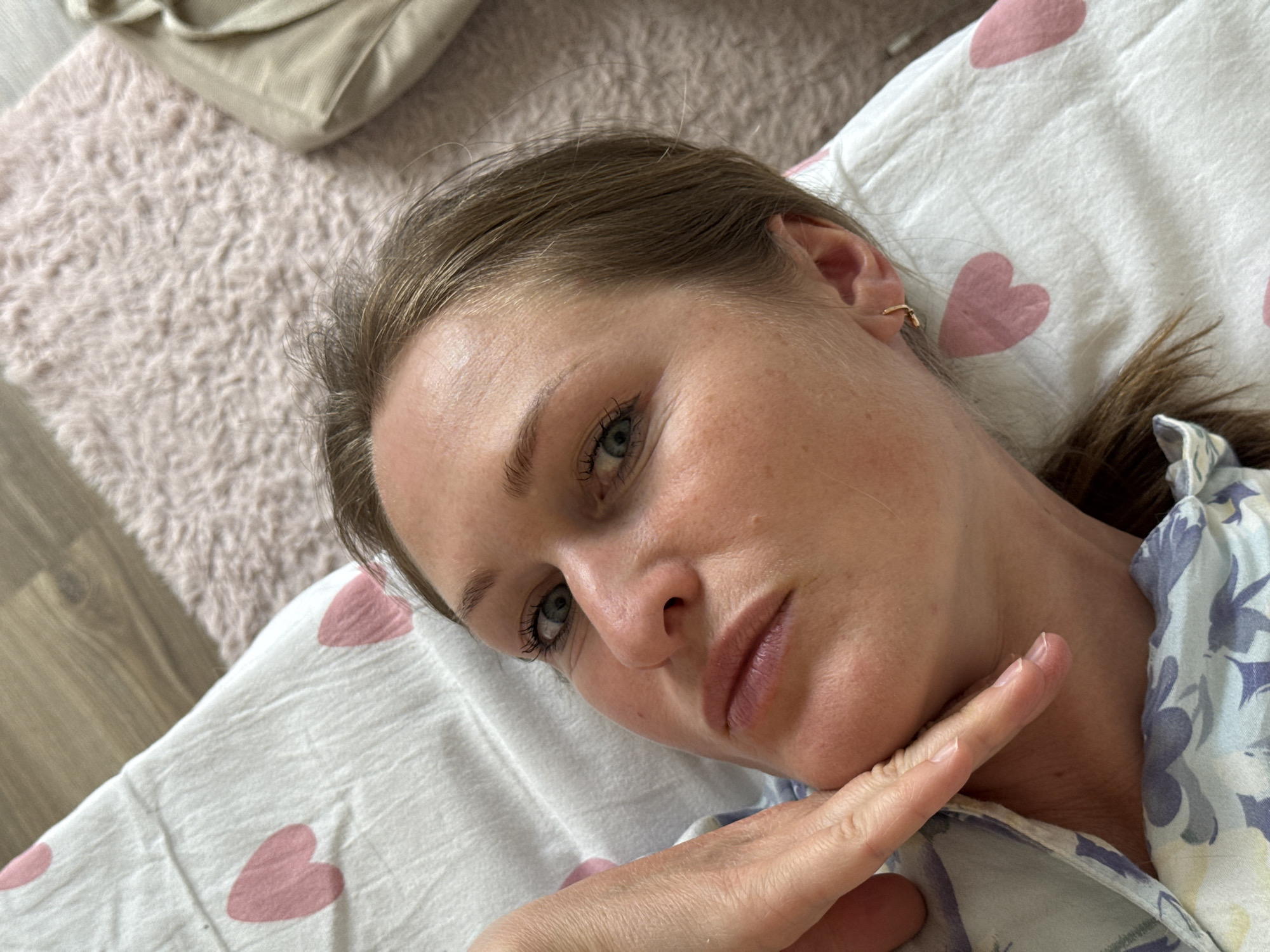 A close-up, slightly tilted shot captures a woman lying down indoors, likely in a bed or on a comfortable surface, in Chaikovsky, Russia. She is positioned on her right side, looking directly towards the camera with a neutral to pensive expression. Her head is gently propped by her right hand, with her fingers resting near her chin.
The woman has light brown hair, pulled back to reveal a small, golden hoop earring in her visible ear. Her eyes are a light blue-green, accentuated by mascara. Her skin appears natural with some visible texture, and her lips have a soft pink hue. She wears a light-colored top or pajama shirt featuring a subtle floral or abstract pattern in shades of blue and purple.
She is resting on a white pillow adorned with scattered pink heart patterns. To the upper left of her, a light pinkish-beige shag rug or fuzzy blanket is visible, with a wooden floor peeking out from underneath. In the top left corner, a beige cushion or pillow can be seen. The lighting is soft and ambient, suggesting an indoor setting without strong direct light, and no text is visible.
