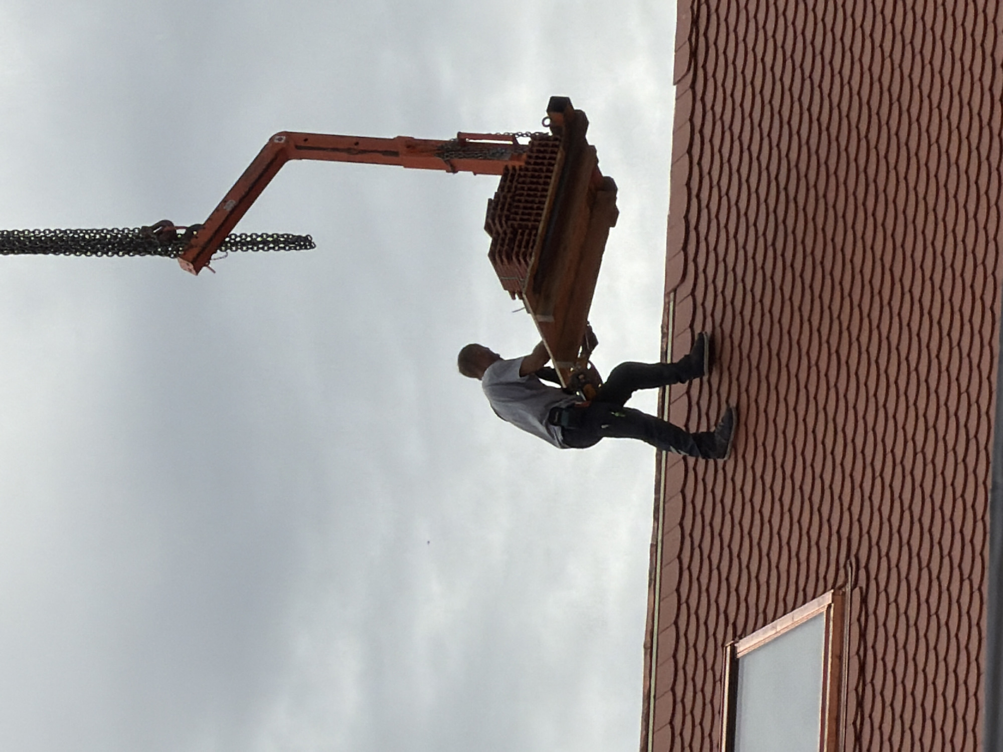 A person wearing a grey shirt, dark pants, and a tool belt stands on a red-tiled roof, holding onto a wooden pallet laden with stacked bricks. The pallet is suspended in the air by an orange crane mechanism and thick chains, being lifted by the crane. The crane arm extends upwards from the right side of the frame. A roof window with a copper frame is visible on the left side of the roof. The sky is overcast and grey. The scene appears to be a construction site in Munich, Germany.