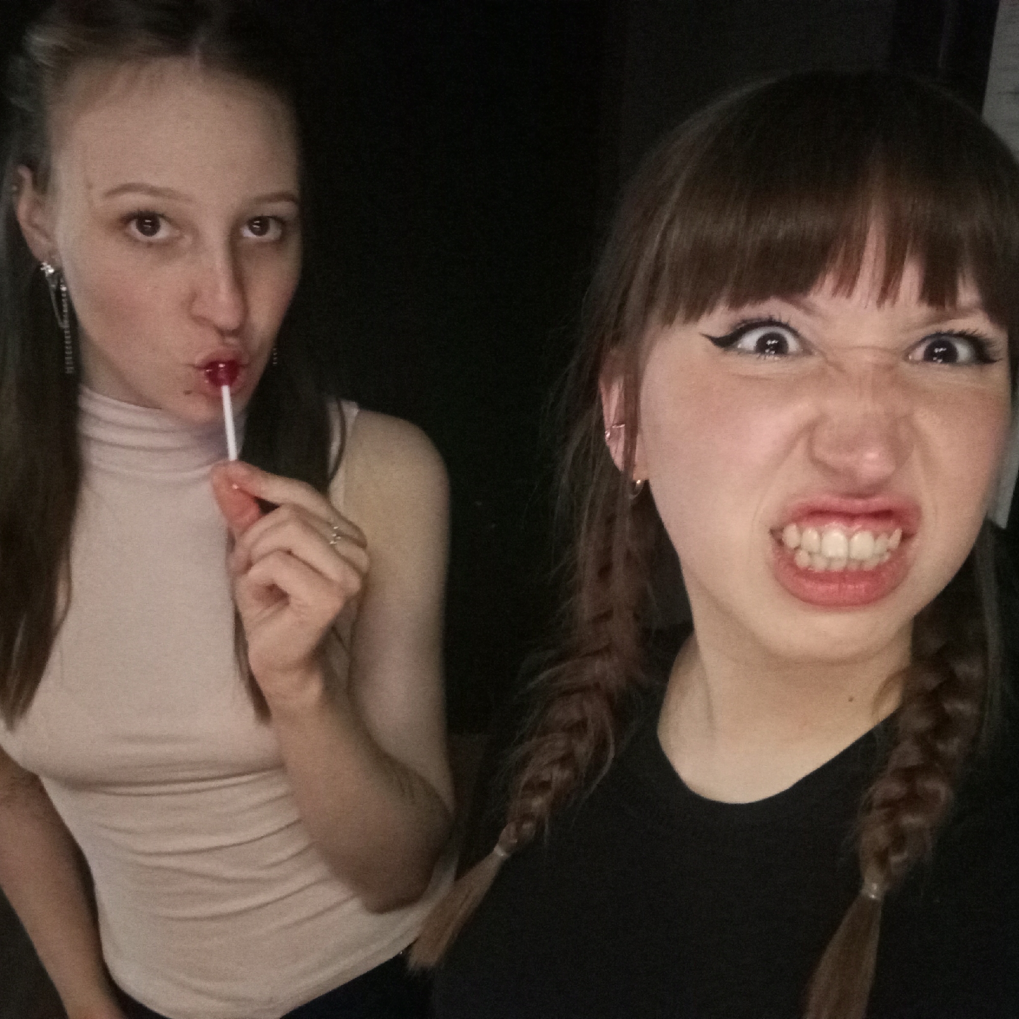 Two young women are posing for a selfie. The woman on the left has long, dark hair, is wearing a light-colored turtleneck shirt, and is holding a red lollipop in her mouth. The woman on the right has dark hair with bangs styled in braids, is wearing a black T-shirt, and is making a scowling face with her teeth bared. The background is dark. There is no indication of the image being taken in Chaikovsky, Russia.