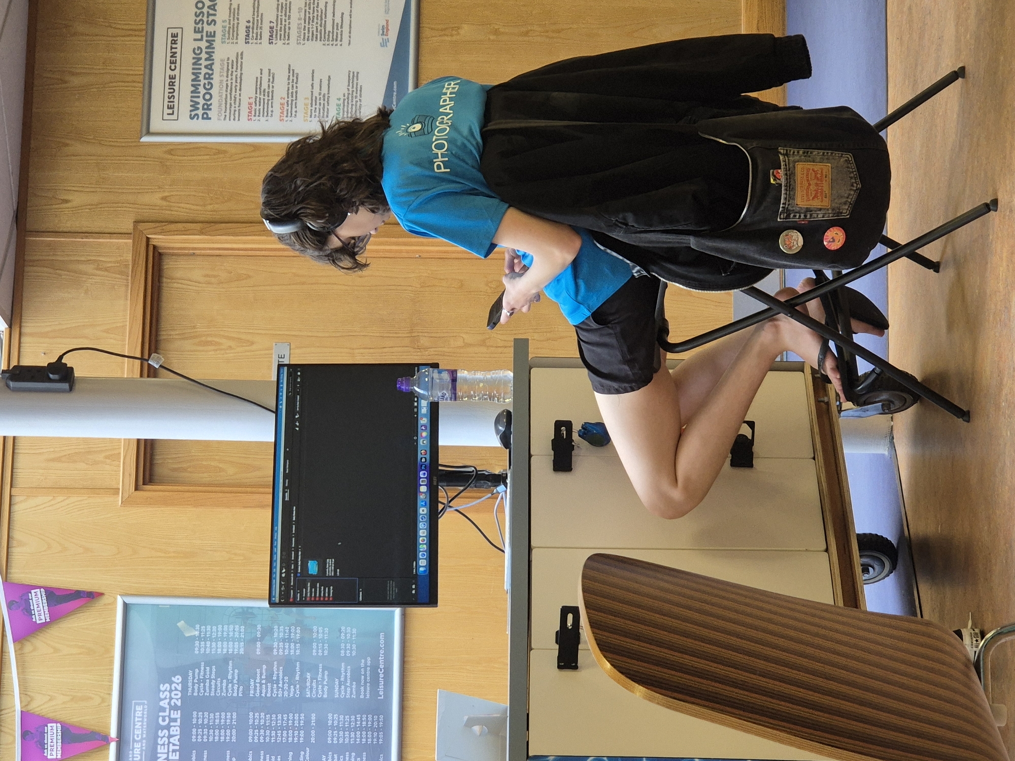 A woman with curly dark hair sits on a black folding chair, facing left, and looking down at a cell phone in her hands. She wears a blue t-shirt with the word "PHOTOGRAPHER" printed on it, along with black shorts and black shoes. She has headphones around her neck and a black jacket hanging on the back of the chair. The chair is positioned next to a white cabinet on wheels. A computer monitor is on the cabinet, displaying a dark screen. A water bottle is also on the cabinet. In front of the cabinet is a desk with a striped wooden top.
Behind the woman and the cabinet, there is wood paneling on the wall. Hanging on the wall are two posters. One has "LEISURE CENTRE" in large letters and the second has "URE CENTRE" and "NESS CLASS" printed on it. Additionally, above the wood paneling, there is another poster with the words "LEISURE CENTRE", "SWIMMING LESSO", and "PROGRAMME STAC" printed on it. The location is the Breckland District in the United Kingdom.
