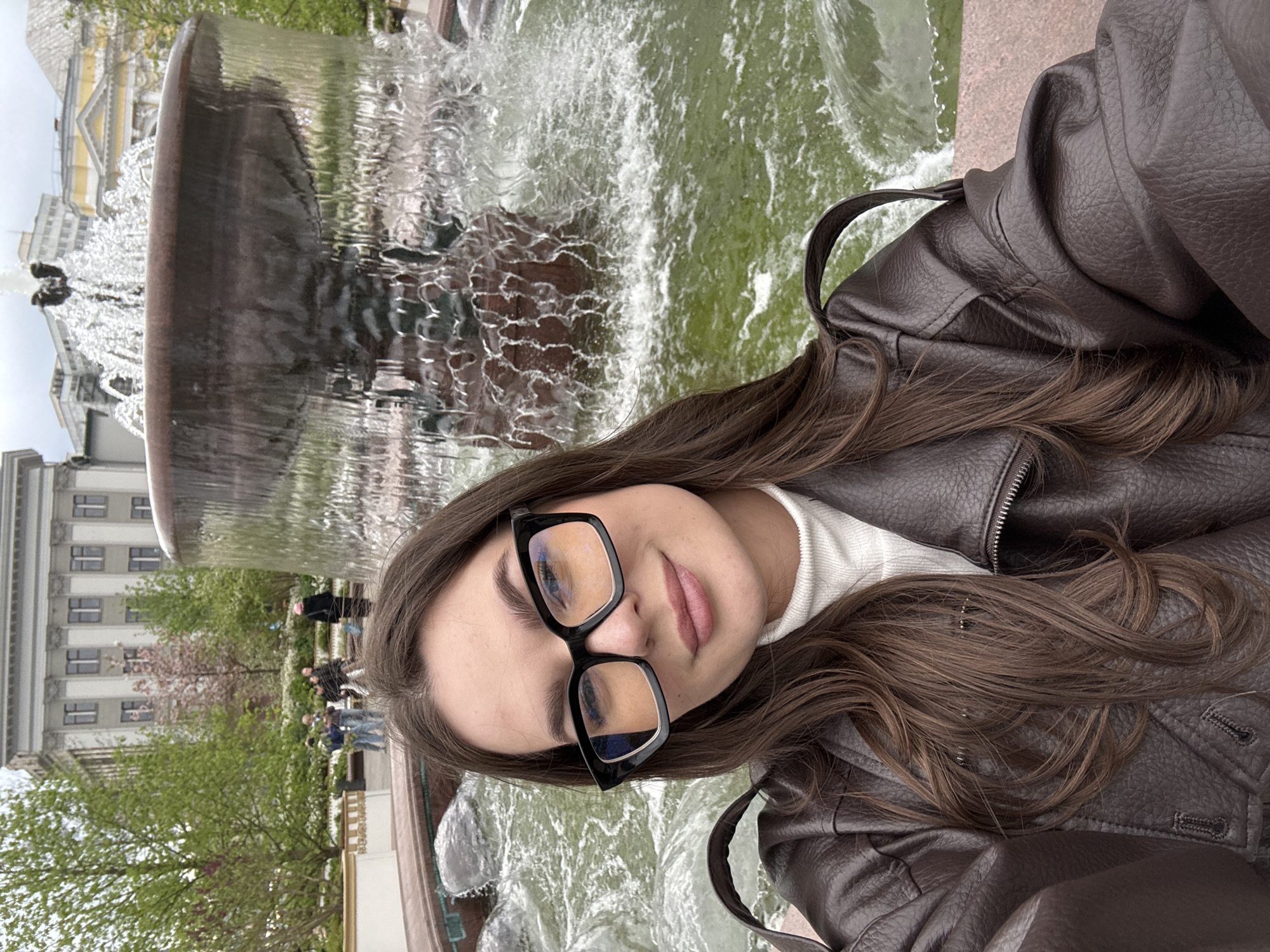A young woman with long, brown hair and wearing black-framed glasses, a white turtleneck sweater, and a brown leather jacket is posing for a selfie in front of a fountain. The fountain is large, with multiple tiers, and water is cascading down. In the background is a building, identifiable as the Moscow State University building in Moscow, Russia. There are other people and trees visible in the background.