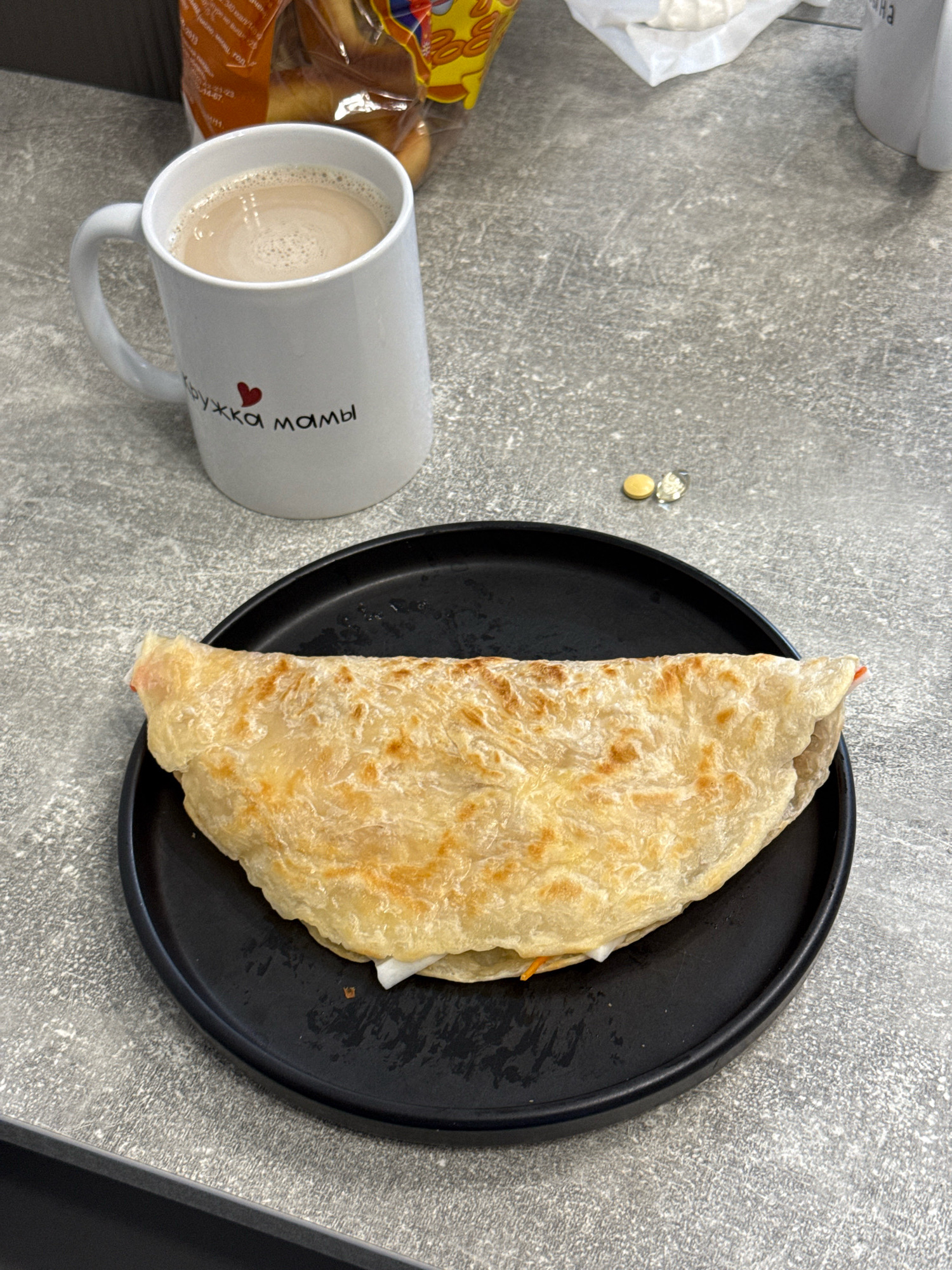A mug of coffee and a folded flatbread stuffed with filling sit on a speckled gray countertop. The white mug has a red heart and black Cyrillic text that reads "кружка мамы" (mom's mug). Next to the mug are two small, round pills. The flatbread is golden brown and appears to be filled with white and orange ingredients. A black plate holds the flatbread. In the background, a bag of baked goods is visible. The scene is likely indoors, possibly in a kitchen or dining area in Lyubertsy, Russia, though no specific landmarks confirm this location.