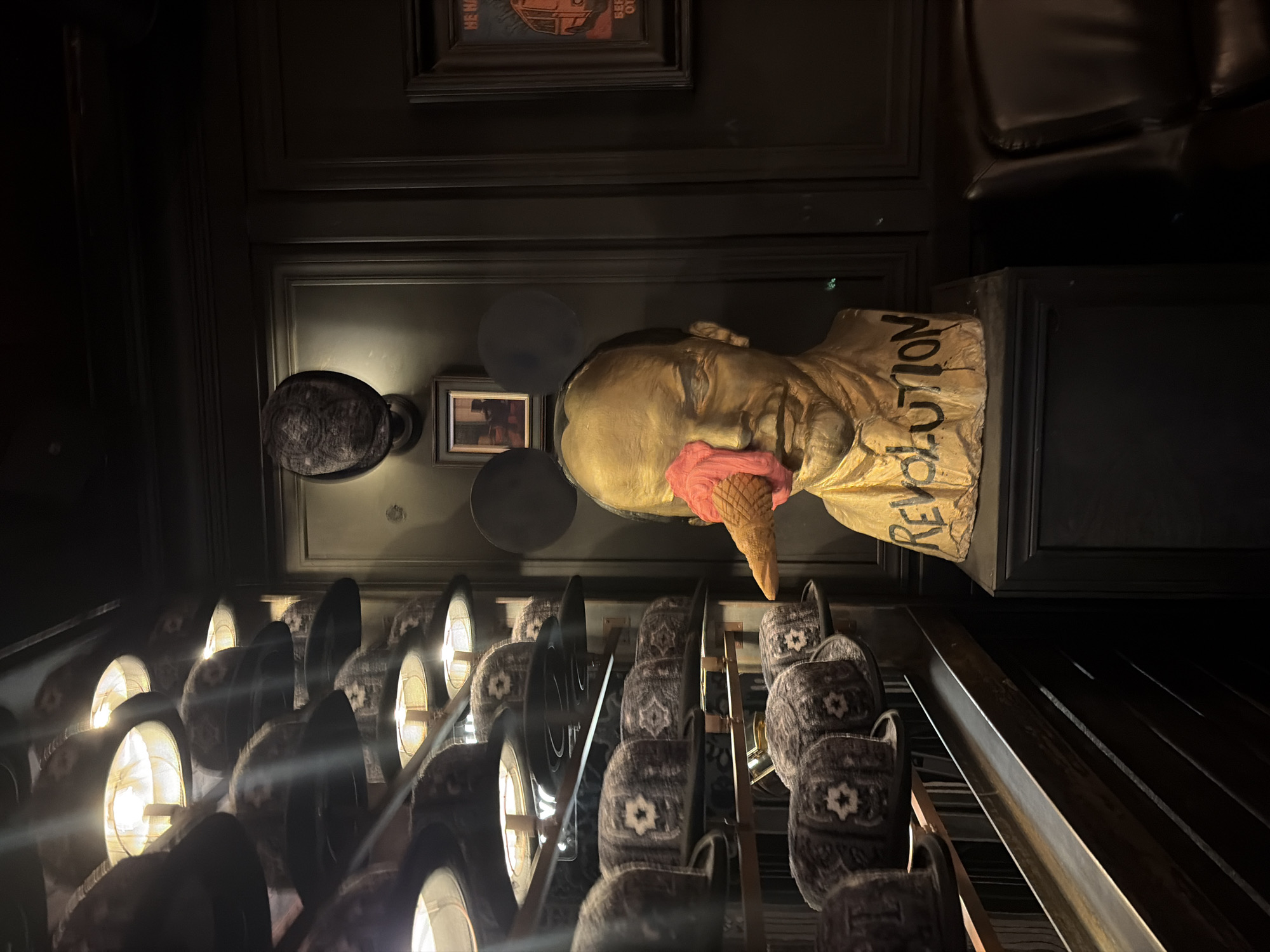 The image shows a dimly lit interior of a shop filled with hats. A golden bust of a person with Mickey Mouse ears and an ice cream cone in their eye is prominently displayed in the center of the frame. The bust has the word "REVOLUTION" scrawled on it. The walls are dark wood paneled, and shelves are lined with various hats, some with ornate patterns and others plain. Lighting fixtures hang from the ceiling, illuminating the hats and the central bust. A framed picture hangs on the wall behind the bust, and another framed poster is visible on the right side of the image. A leather chair is partially visible in the lower right corner. This scene is located in Saint Petersburg, Russia, at the "Hat Museum" (Muzeum Shapok), which is a unique museum dedicated to headwear and is situated in the city center.