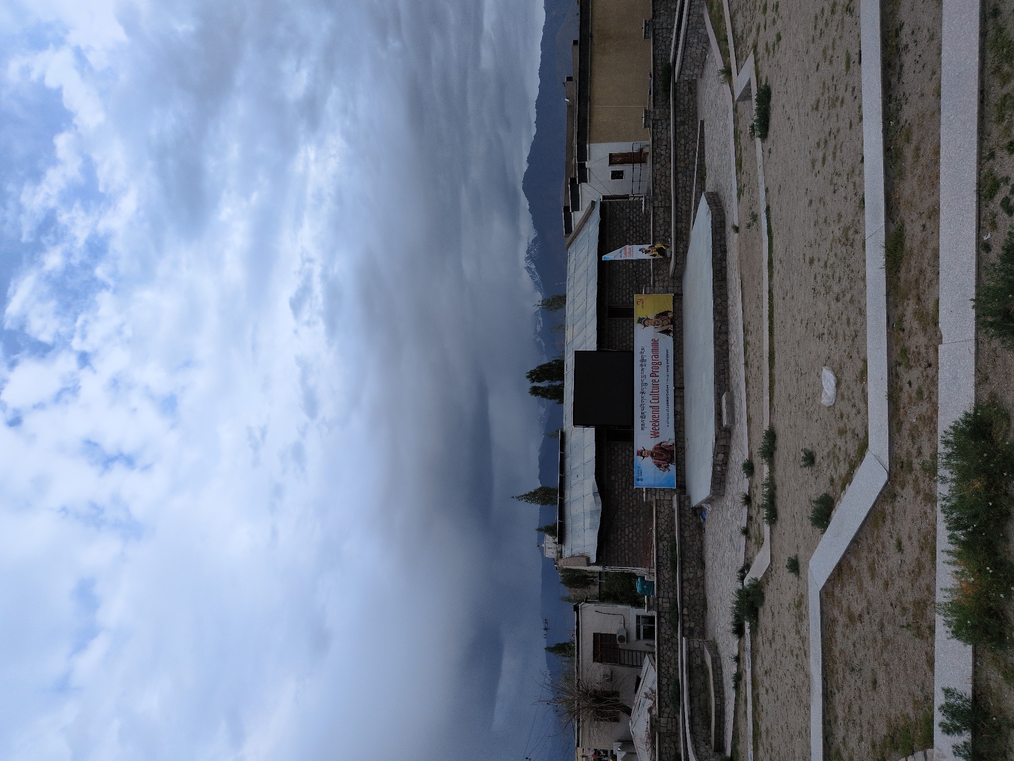 An outdoor amphitheater in Leh, India, is depicted under a cloudy sky. The amphitheater features a circular stage with stone seating areas arranged in tiers. A large banner behind the stage advertises a "Weekend Culture Programme" with images of local people. Surrounding the amphitheater are low stone walls and dry, sandy ground with patches of sparse vegetation. Buildings with corrugated metal roofs and stone walls are visible in the background, along with distant mountains, some capped with snow. The overall scene suggests a cultural event space in a high-altitude desert environment.