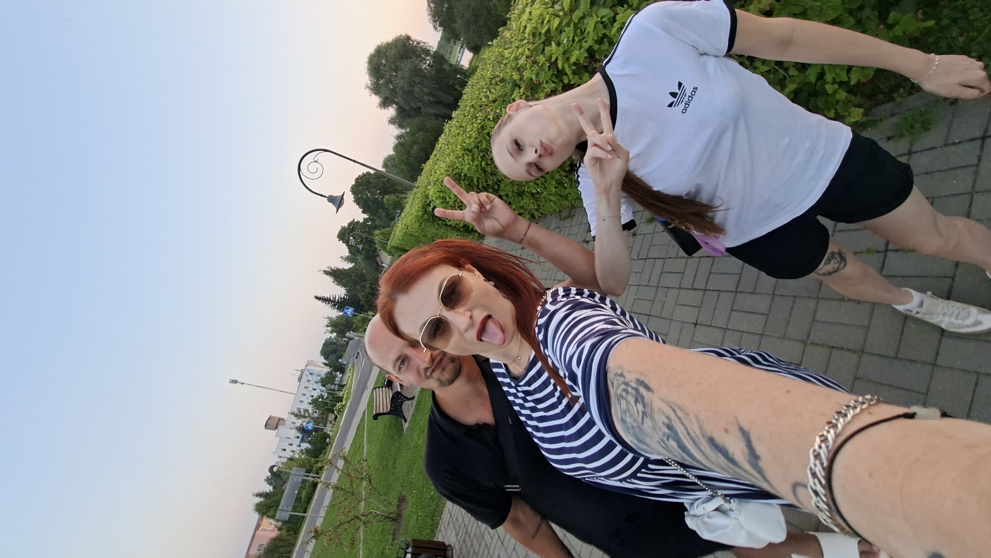 A group of three individuals is captured in a selfie outdoors in Myadzyel, Belarus, during what appears to be late afternoon or early evening, given the soft lighting and sky color. The foreground features the arm of the person taking the selfie, adorned with a silver bracelet and a tattoo. To their left, a bald man in a black t-shirt is partially visible, looking towards the camera with a slight smile. Next to him and slightly in front, a woman with red hair, wearing sunglasses and a striped dress, sticks out her tongue and makes a peace sign with her right hand. To the right of the woman, another individual, likely a young woman, is also making a peace sign with both hands, wearing a white Adidas t-shirt and black shorts.
The setting is a paved walkway lined with a dense green hedge. In the background, a park-like area is visible with trees, a park bench, and a lamppost. Further beyond, buildings and a cell tower suggest an urban or town environment. The overall atmosphere is casual and convivial.
