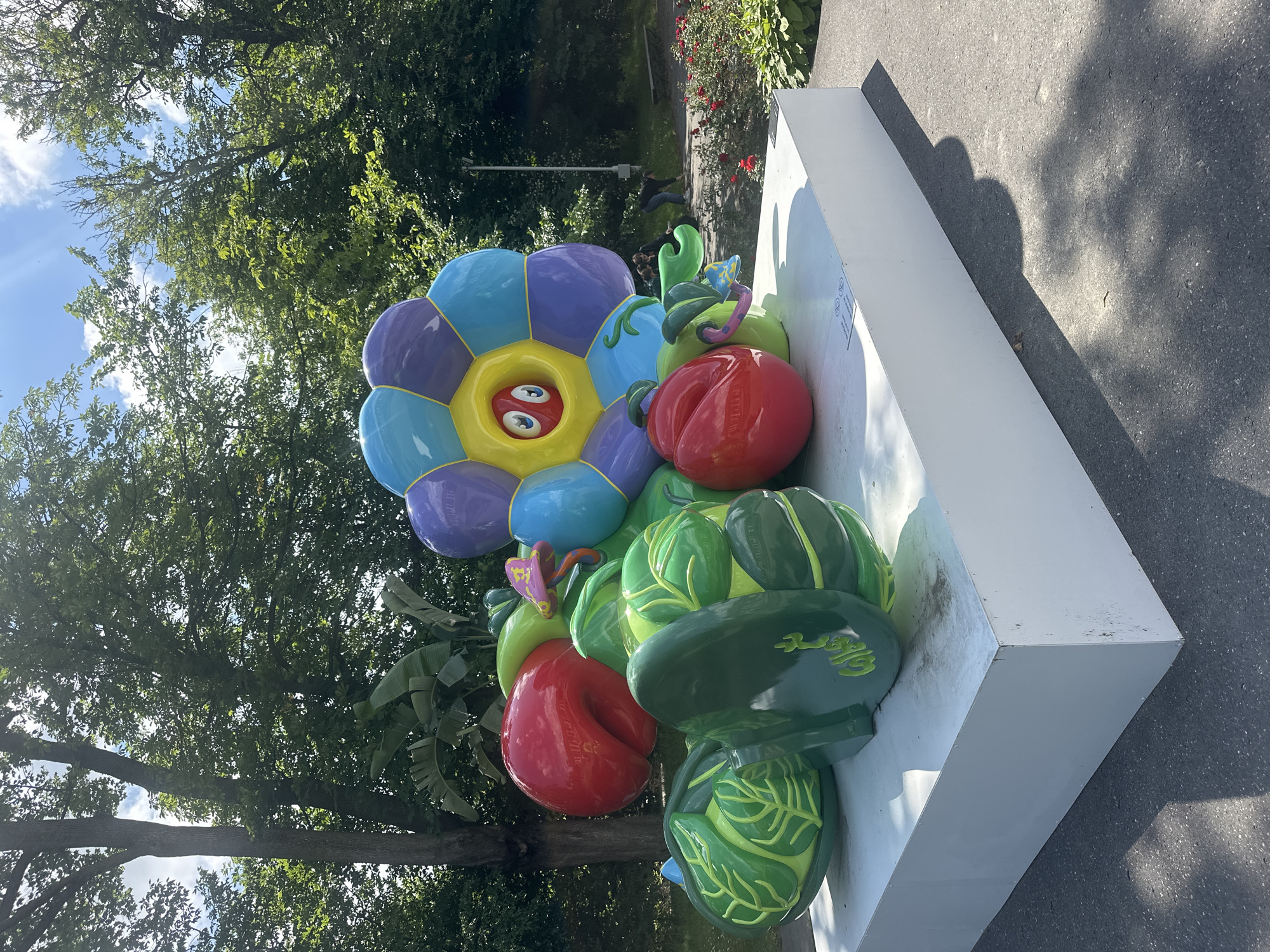 A vibrant, colorful, and whimsical outdoor sculpture resembling a collection of cartoonish vegetables and a flower is displayed on a white pedestal in a park setting. The sculpture features large, brightly colored elements including red apples, green cabbage-like forms, and a large flower with petals in shades of purple, blue, and yellow. The flower has a friendly face with wide eyes and a red center. The sculpture is situated on a paved area in a park with lush green trees and foliage in the background. Several people are visible in the background, some walking and others interacting near a bench. Red rose bushes are also visible in the background. The scene is illuminated by bright sunlight, casting shadows on the ground. The sculpture is located at the Królikarnia Palace, part of the National Museum in Warsaw, Poland, a place known for its sculpture park.