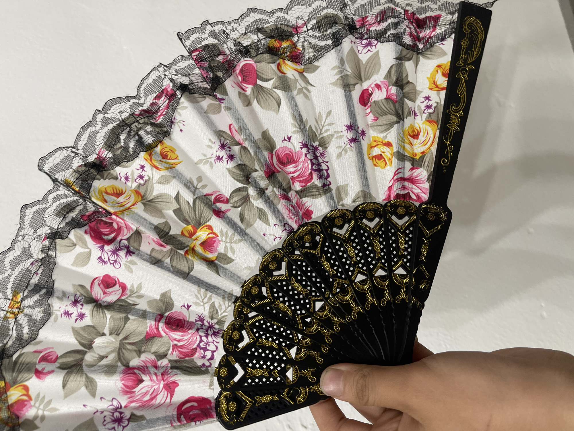 A close-up, eye-level shot shows a hand holding a black, ornate folding fan. The fan is open, revealing a fabric with a white background adorned with a floral pattern of pink, yellow, and purple roses, along with green leaves. Black lace trim with a scalloped edge runs along the outer edge of the fabric. The fan's ribs are black and decorated with intricate gold patterns, featuring geometric shapes and floral motifs. The background is a plain white wall, suggesting an indoor setting. No specific location in Bishkek City, Kyrgyzstan, is visible.