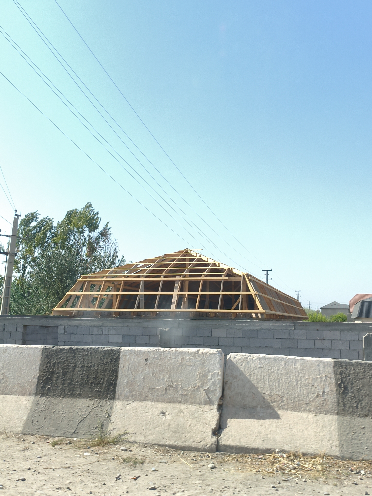 A construction site in Boghparvar, Tajikistan is captured from a low vantage point. In the foreground, two concrete barriers, one with a faded black and white pattern, line a dusty ground covered with scattered pebbles and dry grass. Behind the barriers, a wall constructed from grey concrete blocks is visible. Rising above this wall is the wooden frame of a uniquely shaped building, possibly a geodesic dome or pyramid. The structure is incomplete, with exposed wooden beams and no roofing material yet in place. To the left of the building, a tall, weathered utility pole stands with electrical wires stretching across the clear blue sky. Behind the utility pole and to the left of the building, a dense cluster of green trees sways in the wind. In the distance, several houses with tiled roofs are visible, along with more utility poles, suggesting a semi-urban or suburban setting. The overall impression is one of ongoing construction under a bright, sunny sky.