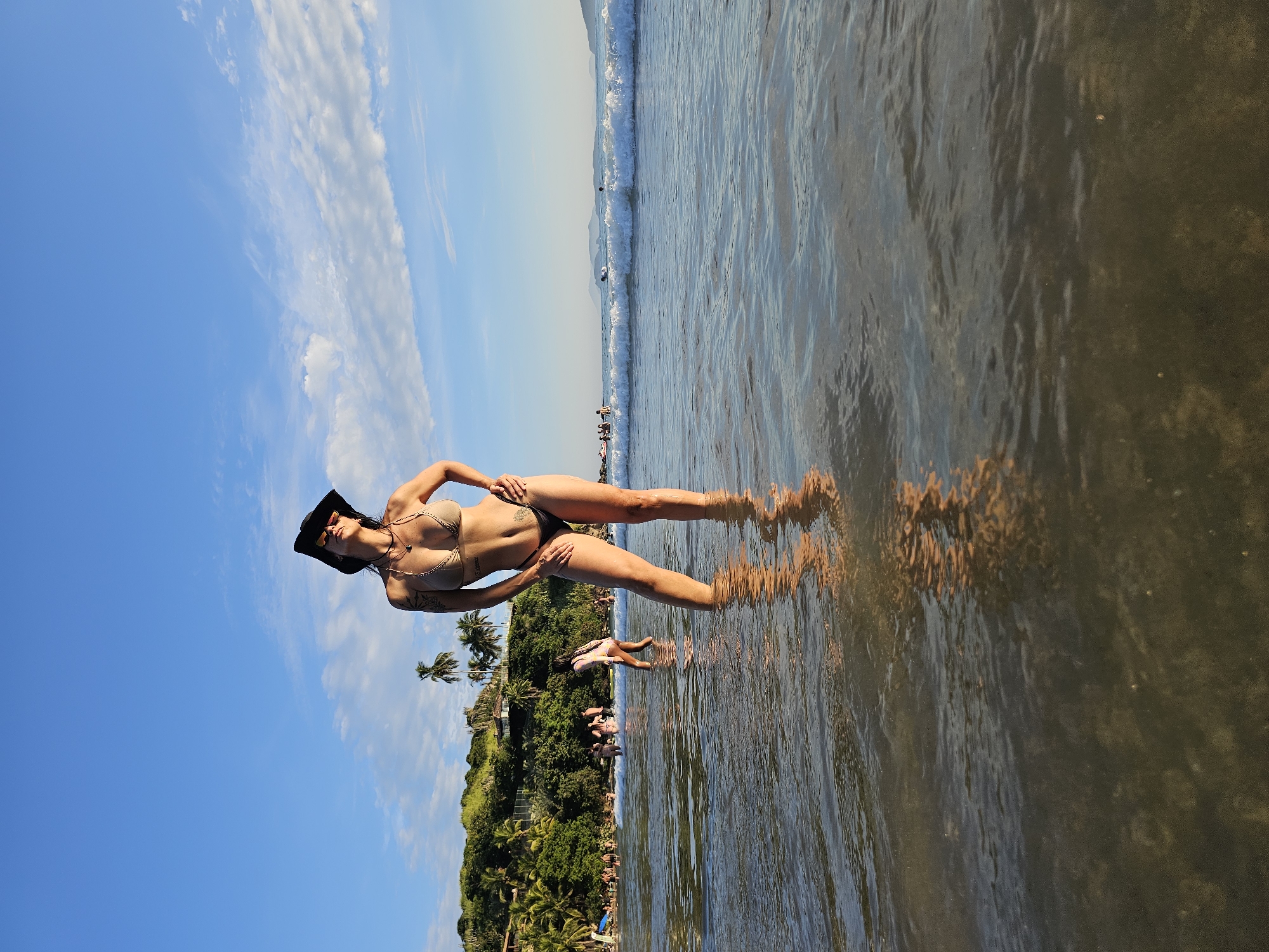 A woman in a bikini and a black hat is standing in the water at a beach. She is posing for the camera with her hands on her hips and her legs spread apart. The water is clear and shallow, and the sand is a light tan color. In the background, there is a lush green hillside and a blue sky with white clouds. People are visible on the shoreline in the distance. The location is Armação dos Búzios, Brazil.
