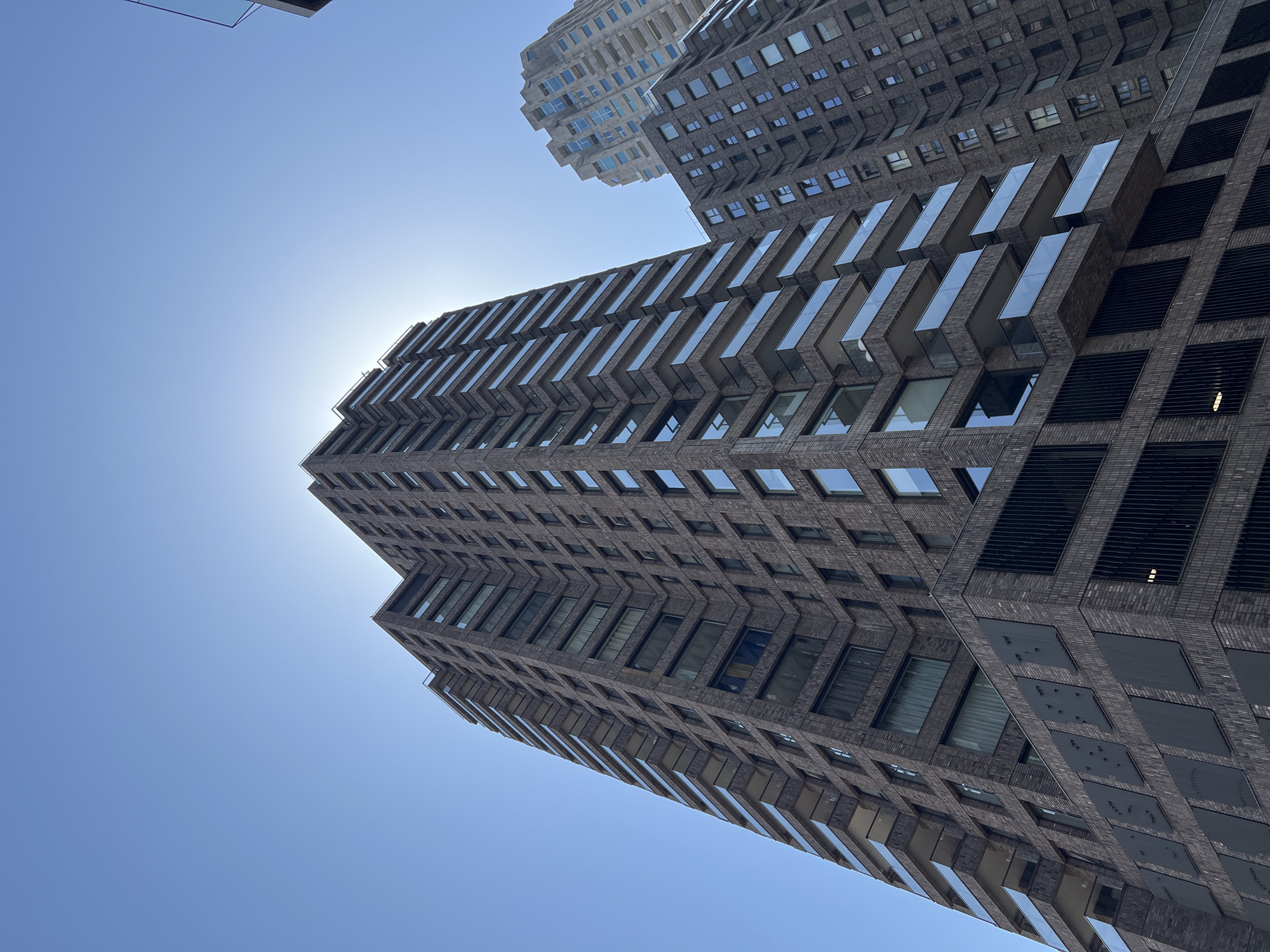 In Rotterdam, Netherlands, a low-angle shot captures the imposing facades of modern high-rise apartment buildings against a clear blue sky. The building in the foreground, constructed with dark brick, features a striking design with numerous balconies extending outwards, each enclosed by clear glass railings. These balconies are staggered, creating a dynamic visual rhythm on the building's exterior. The lower section of this building is characterized by large, dark rectangular panels, some with textured surfaces and others with vertical slats, suggesting potential access points or ventilation systems.
To the right and slightly behind, another tall building is visible, its facade constructed from a lighter-colored material, possibly concrete. This building also has many windows and balconies, but its architectural style appears more angular and stepped, with prominent recessed sections that house balconies. The sunlight is intense, creating a bright halo effect around the top of the foreground building, hinting at the sun's position behind it. The overall scene is one of urban density and contemporary architecture, with no people or vehicles immediately discernible.