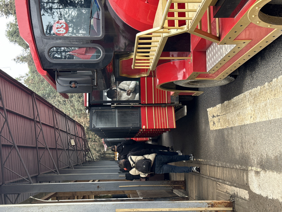 An outdoor scene depicts a tourist shuttle stop in Wulong District, China, characterized by a forested background under an overcast sky. A multi-carriage, red and black tourist shuttle, resembling a stylized train with gold trim and steps, is parked parallel to a covered platform. The lead vehicle displays a "03" designation in a red circle on its front section. Several passenger doors on the carriages are open.
A minimum of four individuals are present. One person, wearing a light-colored puffer jacket, is seated inside a shuttle carriage and is actively observing a mobile phone. On the adjacent platform, three individuals are observed walking away from the camera, consistent with boarding or disembarking activity. The individual closest to the camera on the platform wears a beige puffer jacket and a dark backpack.
The platform features a wooden plank walkway and is covered by a reddish, corrugated metal roof supported by dark metal trusses and posts. The ground surface for the shuttle consists of asphalt with a faded yellow painted line. Evergreen trees are visible behind the platform and shuttle, indicating a natural, possibly mountainous, environment.