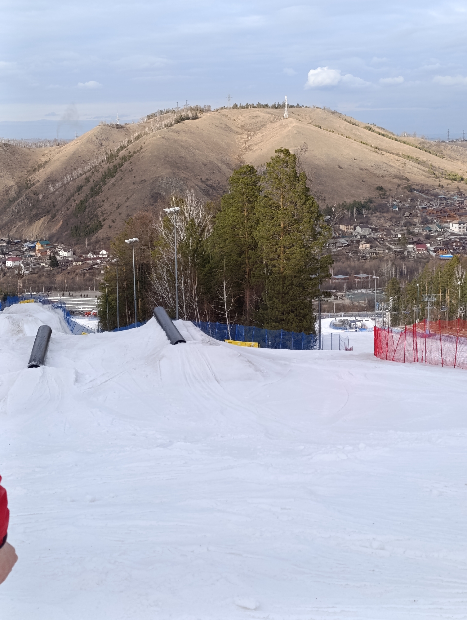 The image depicts a snow-covered ski slope, likely a terrain park, in Krasnoyarsk, Russia, with a hill in the background topped with a cellular tower. Several man-made features are present, including two parallel metal rails on small snow ramps, suggestive of a jump. Blue and red safety netting is visible along the sides of the slope. A village or urban area sits at the base of the hill behind the ski slope. The sky is overcast. There is a glimpse of a person wearing a red jacket on the left side of the image.
