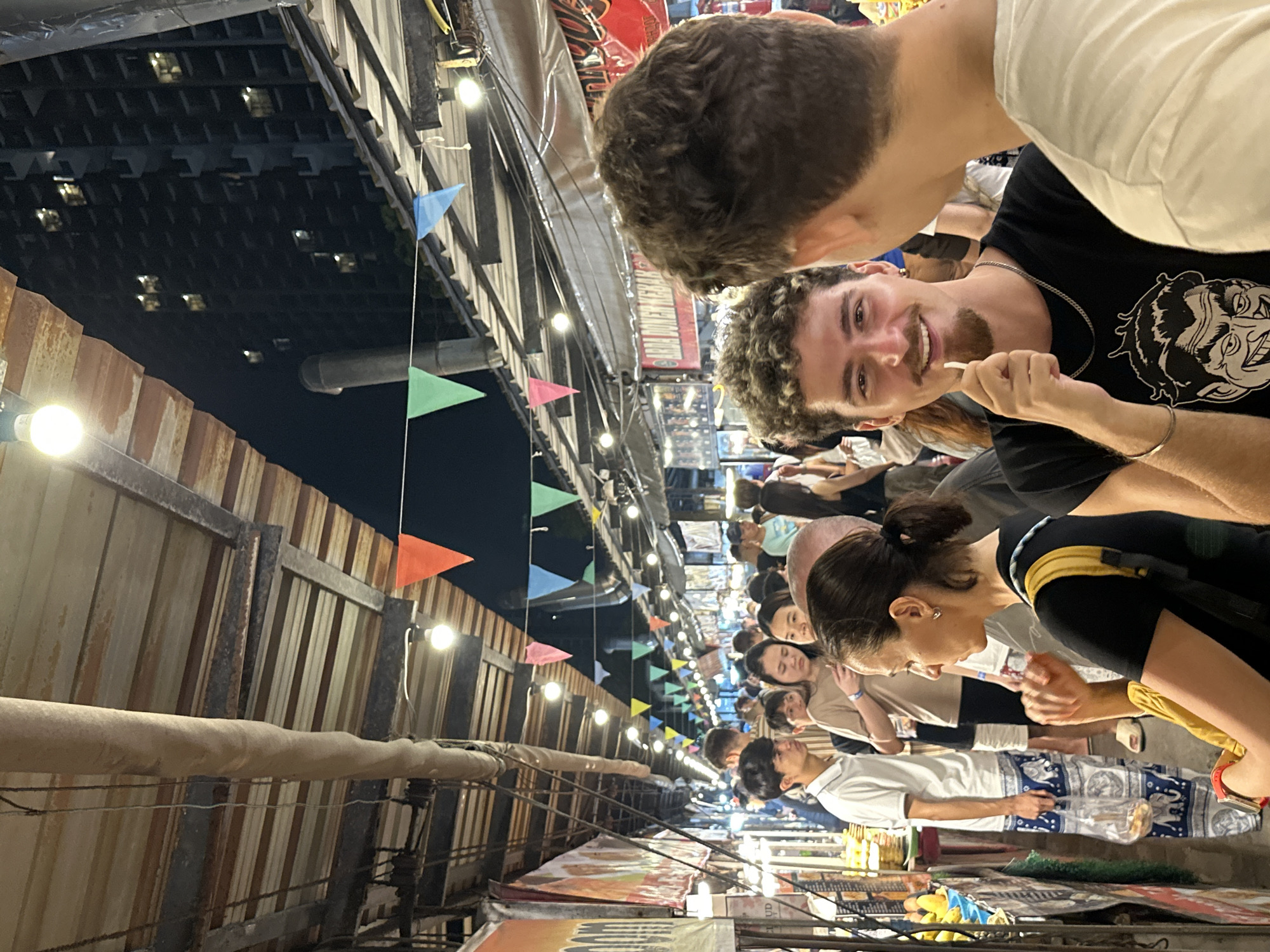 This photo was taken in Pattaya City, Thailand. The scene captures a bustling night market with numerous people browsing the stalls. Several overhead string lights and colorful triangular flags decorate the market. The foreground features a person holding a toothpick in their mouth, while another person with short brown hair stands close by. Many other people are visible further down the crowded aisle, browsing the market stalls. These stalls display various goods. The market appears to be set up between corrugated metal walls, with a low roof covering the area, the ceiling is visible on the left of the image and black sky is visible on the top left.