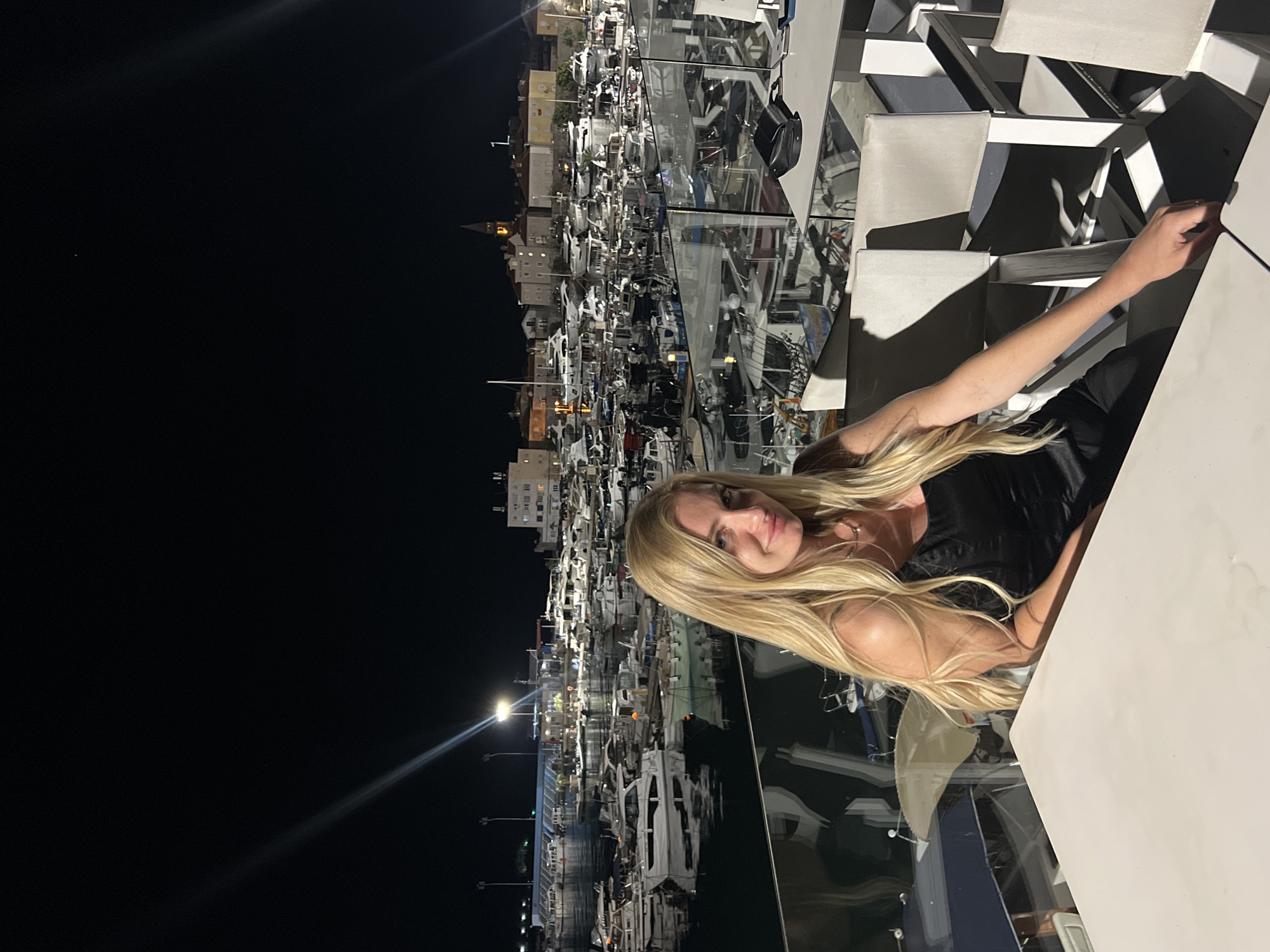 A woman with long blonde hair is sitting at a table at a restaurant in Budva, Montenegro. The background shows the city's marina, filled with boats, illuminated at night. The restaurant has white tables and chairs. The woman is wearing a black top. Her arm is extended towards the table.