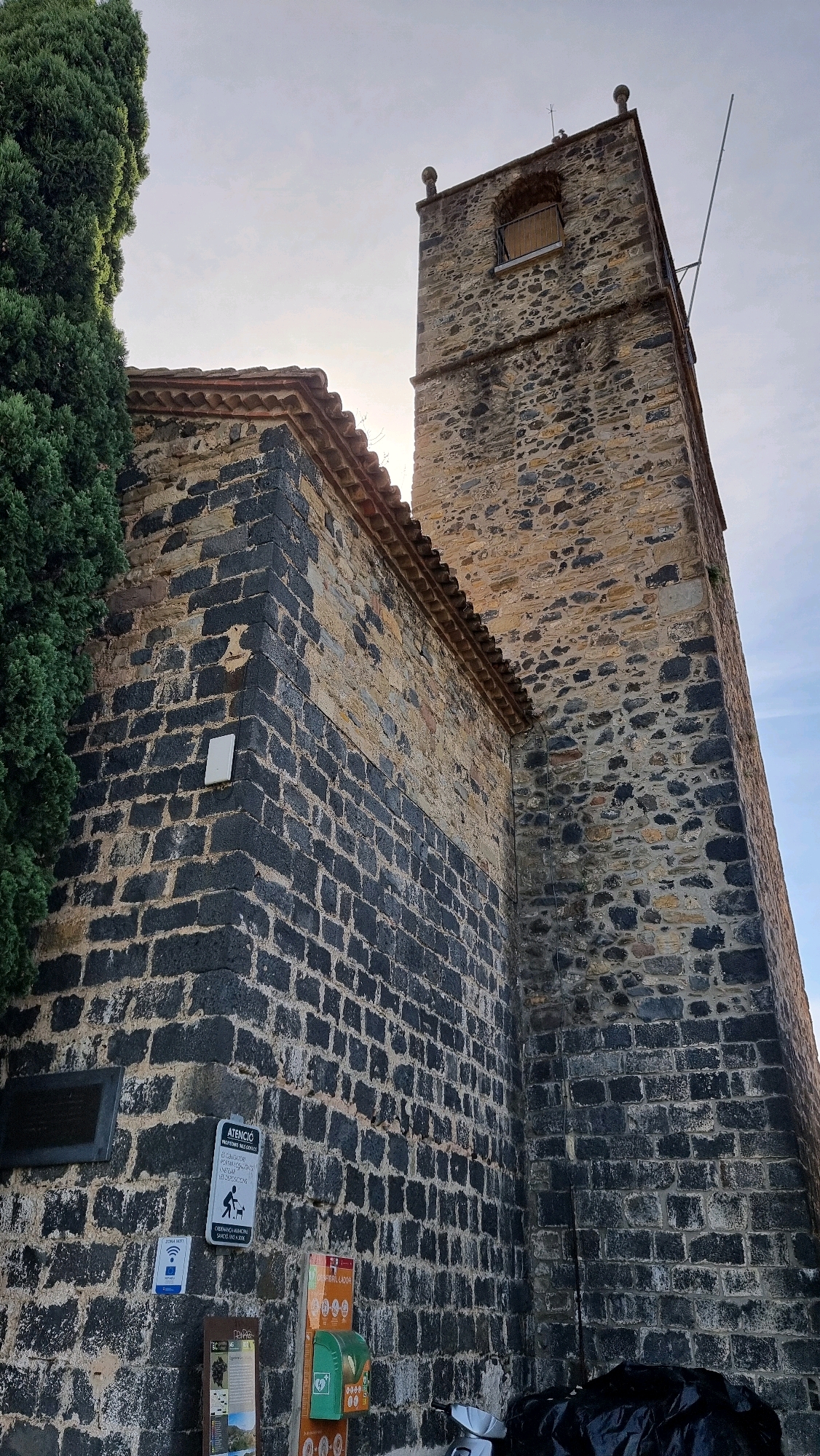 This image captures a view of the Bell Tower of the Church of Sant Miquel in Castellfollit de la Roca, Spain. The tower is constructed of dark, roughly hewn stone blocks with lighter stone interspersed. It has a single arched opening near the top, covered by a wooden louvered shutter. Two decorative finials crown the top corners of the tower. A long, thin metal pole extends from the right side of the tower.
Adjacent to the tower, on the left, is a section of the church building. Its lower portion is made of the same dark stone as the tower, while the upper portion is constructed of lighter-colored stone. A terracotta tiled roof with a decorative edge caps this section. A large, dark green coniferous tree is visible to the far left, partially obscuring the church building.
At the base of the church wall, several items are mounted:
* A dark rectangular plaque, possibly a historical marker.
* A white rectangular object.
* A sign with text and a graphic of a person with a dog, indicating rules or information about pets.
* A blue and white sign with Wi-Fi symbols.
* A tall vertical sign with text and graphics.
* An orange and green public access defibrillator unit.
Below these, a white scooter and a black tarp-covered object are partially visible on the ground. The sky is overcast, with a bright, diffused light suggesting it is either early morning or late afternoon. The overall scene depicts a historical religious building within a Spanish town.