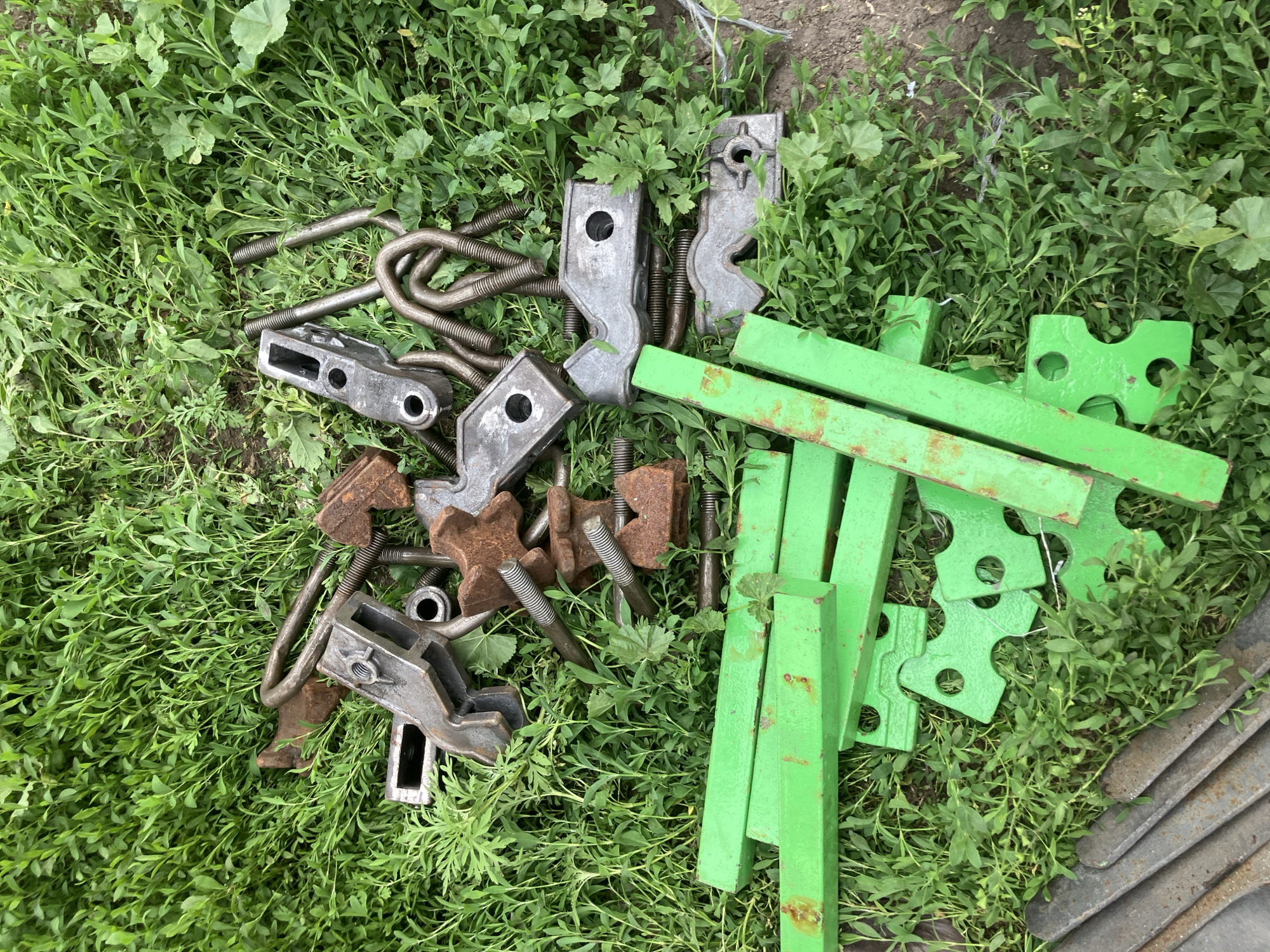 In Vovkivka, Ukraine, a collection of metal hardware is scattered across a patch of green grass. The items include several U-bolts, some of which are rusted, and various clamps and brackets, also showing signs of rust. Interspersed with these are several bright green metal bars and shaped pieces, some with circular cutouts. The scene is outdoors, with the vegetation appearing dense and somewhat overgrown.