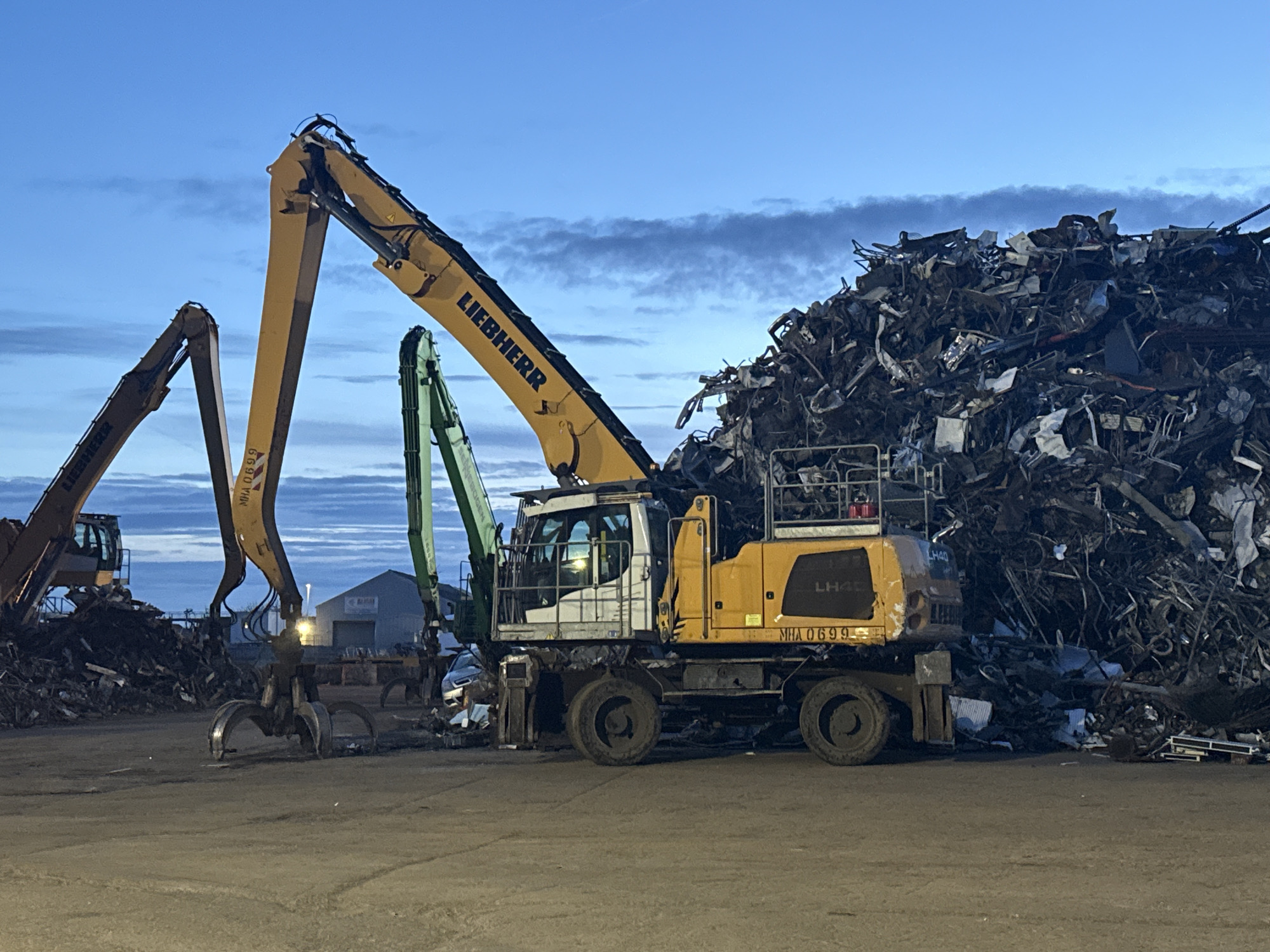 The image shows a scrap metal yard in Gorleston-on-Sea, United Kingdom at dusk. Dominating the scene is a large pile of scrap metal, including what appears to be crushed vehicles and assorted metallic debris. Two large Liebherr hydraulic material handlers are visible. One, prominently positioned in the foreground, is yellow and white with the model number LH40 and the identifier "MHA 0699" displayed on the side. Its grapple attachment is lowered. Another similar material handler is visible further in the background. In the distance, a building with a sign is partially visible. The ground is paved and appears to be dirtied by the scrap processing activity. The sky is a blend of blue and grey, suggesting twilight.