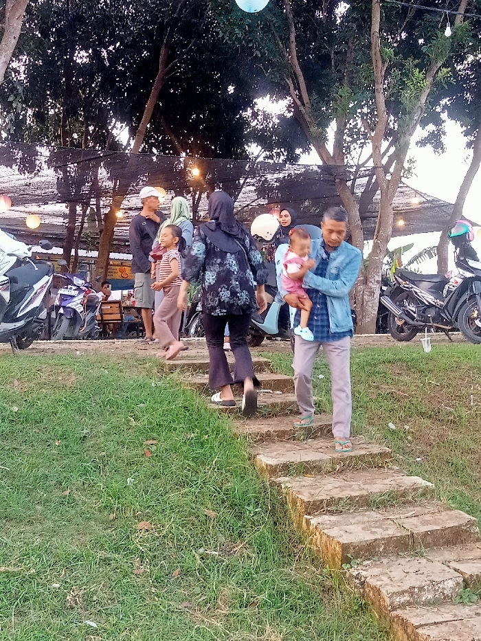 The image shows a group of people walking down a set of concrete steps in a park located in the city of Jakenan, Indonesia, at coordinates -6.758 latitude and 111.075 longitude. There are several trees in the background. Two of the people are wearing traditional clothing, one is holding a small child, one is wearing sandals and one is wearing a blue jean jacket. The person wearing the blue jean jacket is walking down the steps. The people are walking from left to right. The background includes a black plastic fence with lights, and two motorcycles. The ground is covered in green grass. There is a large tree to the left of the steps. The image was captured on a partly cloudy day. There are lights strung up in the background. The people appear to be in a park or a public area. The ground is covered in green grass. The image was taken at an outdoor location.