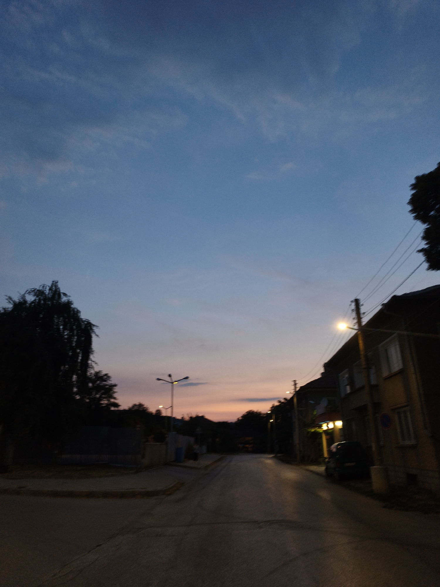 The image captures a street scene at dusk in Lovech, Bulgaria. The sky is a gradient of deep blue fading into hues of orange and pink near the horizon, with wispy clouds scattered throughout. Silhouetted against the sky are the dark forms of trees on the left and the rooftops of buildings on the right. A streetlamp illuminates the road, casting a warm glow. The road itself appears to be paved and wet, reflecting the light. Several houses with visible windows line the street. A dark-colored car is parked on the right side of the road, and a few more streetlights are visible in the distance, stretching down the street. The overall atmosphere is quiet and still, typical of a residential area at the end of the day.