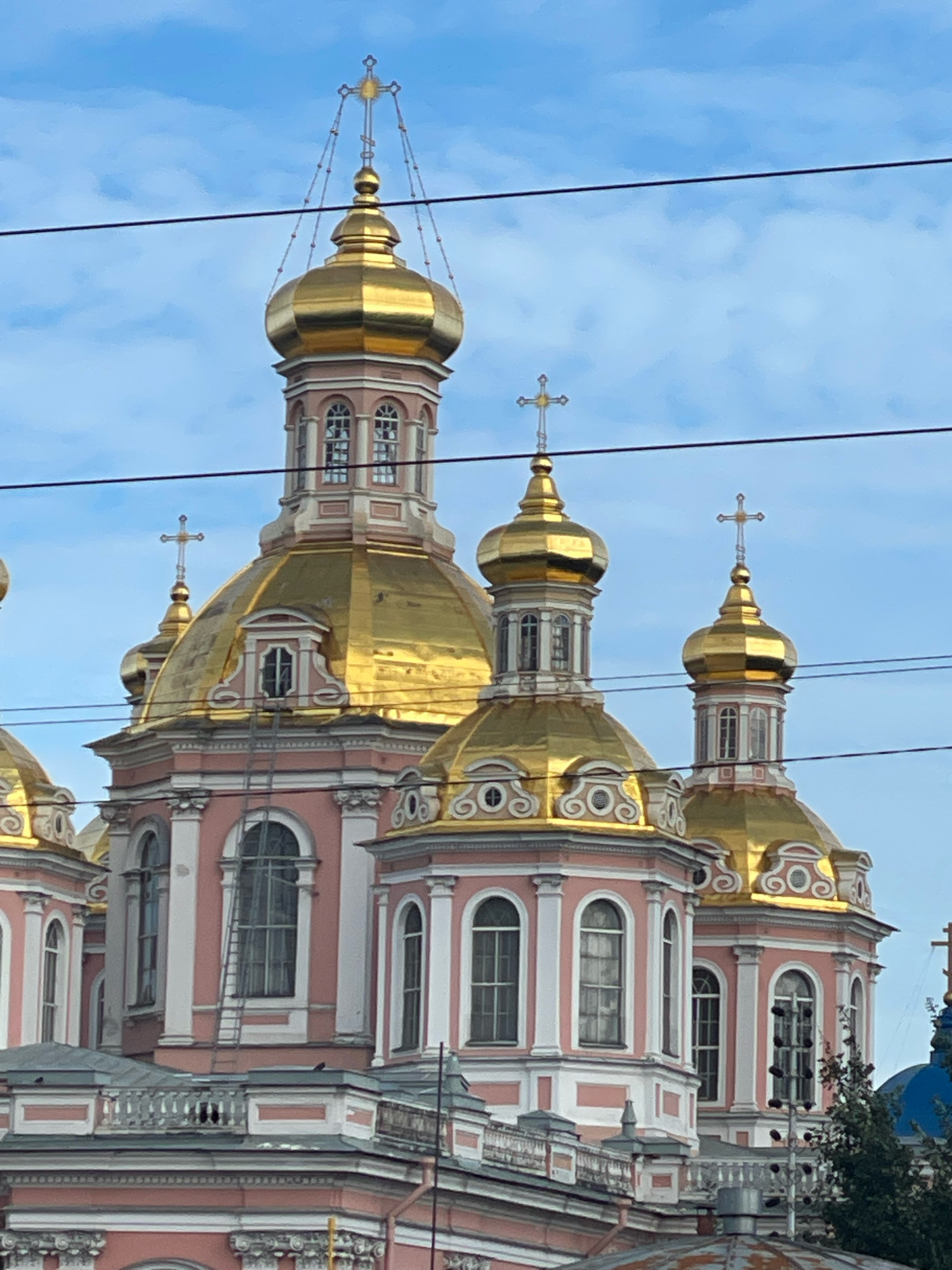 The image shows a close-up view of several golden onion domes and pink and white architectural elements of a church in Saint Petersburg, Russia. The church has multiple levels with arched windows and decorative white trim. Several wires are visible in the foreground, running horizontally across the frame, obscuring parts of the sky and the upper portions of the domes. The sky is partly cloudy with a pale blue hue. In the bottom right corner, a portion of a blue dome and some green foliage are visible, suggesting it is part of a larger complex or neighboring building. There are no people or discernible actions depicted in the image. The overall scene captures the ornate and characteristic architecture of Orthodox churches in Saint Petersburg.