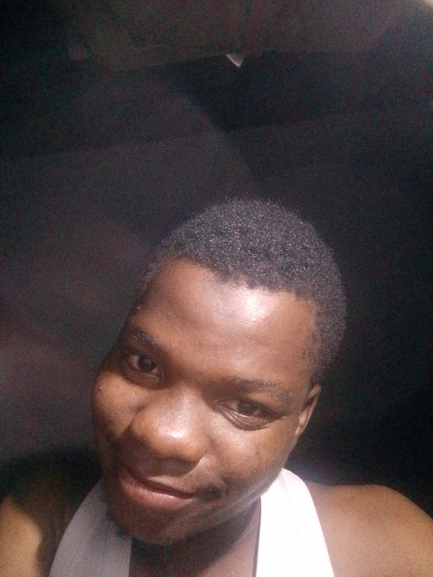 A selfie of a dark-skinned person with short, dark hair wearing a white tank top. The background is dark and out of focus, suggesting an indoor location. The location is Unknown, Unknown.