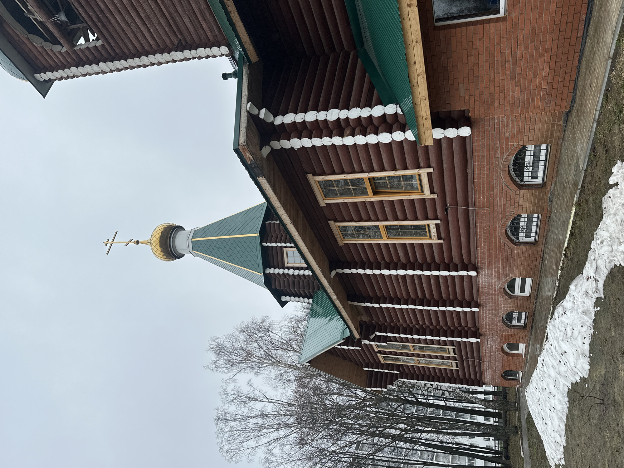 The image depicts a distinctive religious building, likely an Orthodox church or chapel, set against an overcast sky in Chaikovsky, Russia. The building's architecture is a blend of wooden log construction and a brick foundation.
The main structure features dark brown, horizontally laid wooden logs, adorned with decorative white-painted, scalloped ends along the eaves and corners. Rectangular windows with grid patterns and light-colored frames are visible within the wooden sections. The roofs are a vibrant green, made of corrugated metal. A prominent golden onion dome, topped with a golden Orthodox cross, rises towards the left side of the frame.
The lower portion of the building consists of a red brick foundation, which includes several arched windows, some fitted with decorative black metal grilles. Adjacent to the building, a narrow paved path runs alongside, with visible patches of melting snow on the ground, indicating a late winter or early spring season. Bare deciduous trees stand in the background, partially obscuring what appear to be apartment buildings. The sky is uniformly grey and overcast. No specific activity is occurring, and there is no visible text in the image. The scene conveys a tranquil, muted atmosphere characteristic of the transitional seasons.