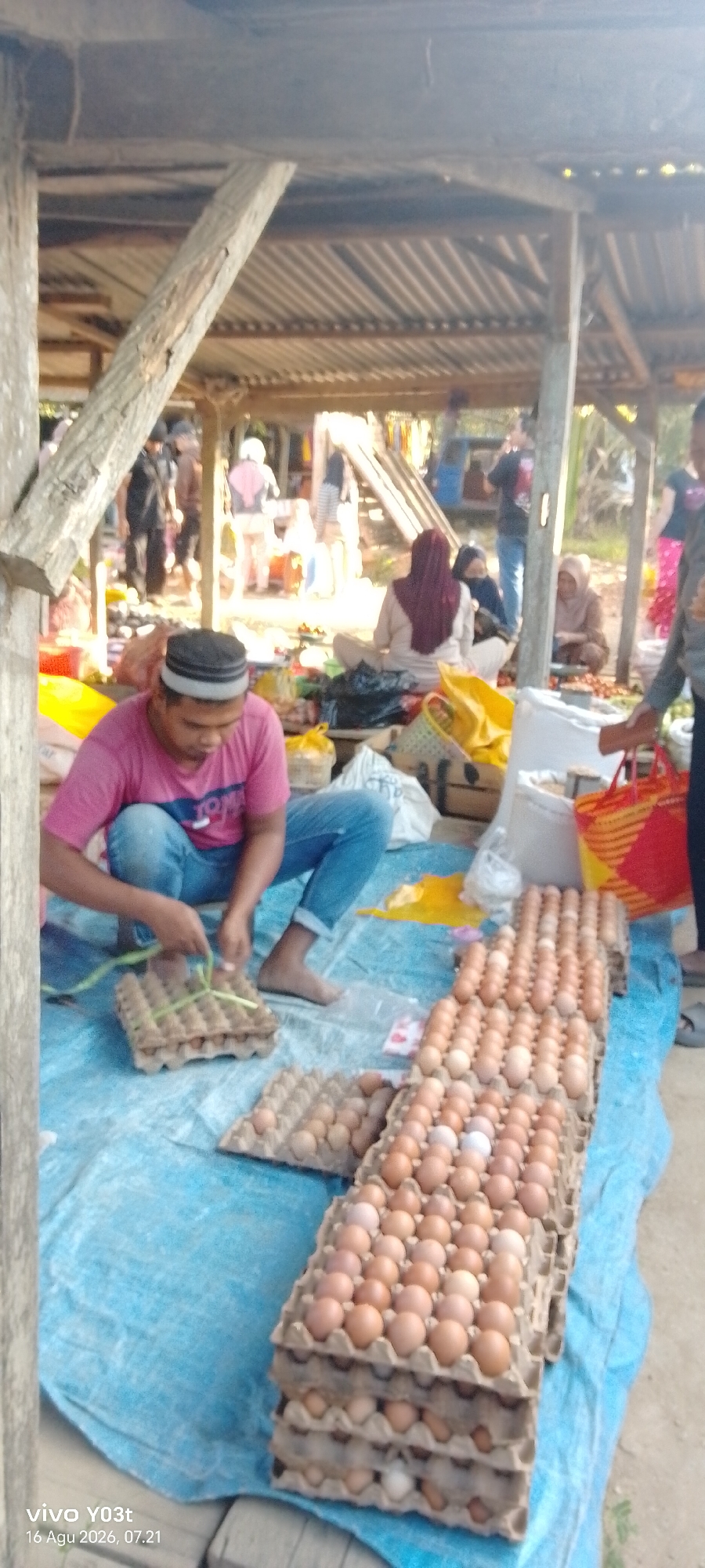 The image depicts an outdoor market scene in Palosilae, Indonesia. In the foreground, a man wearing a pink t-shirt and blue jeans is sitting on a blue tarp, sorting or packaging eggs from cardboard cartons. He is holding a green string and appears to be tying it around a carton. Several stacks of egg cartons, filled with brown and white eggs, are arranged on the tarp in front of him. In the background, other market-goers are visible. Some are sitting or standing around stalls with various goods, possibly food items, displayed on the ground. The market appears to be under a simple, open-air structure with a corrugated metal roof supported by wooden beams. The overall environment suggests a bustling local market.