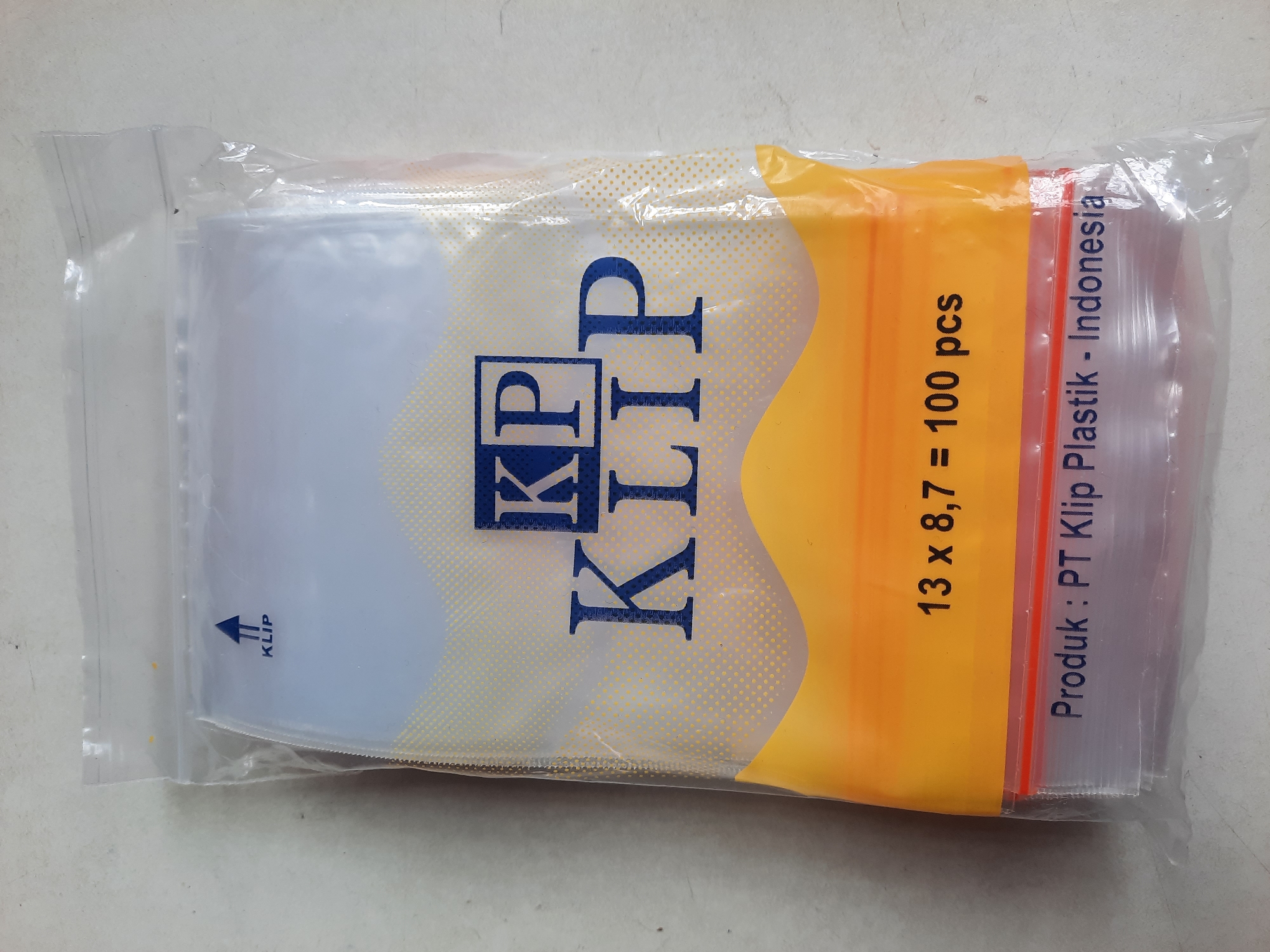 The image shows a clear plastic bag containing multiple small, empty plastic bags. The outer packaging of the bag is also clear plastic with a ziplock closure at the top. A blue and yellow design is printed on the front of the bag. This design includes the text "KP KLIP" in large blue letters, and below it "13 x 8,7 = 100 pcs". At the bottom of the bag, the text "Produk : PT Klip Plastik - Indonesia" is printed in dark blue. The bag is placed on a light-colored, textured surface, possibly a table or floor. There are no people or animals visible in the image. The scene is indoors, and the lighting appears to be neutral. There is no information in the image that suggests a specific location in Lhokseumawe, Indonesia.