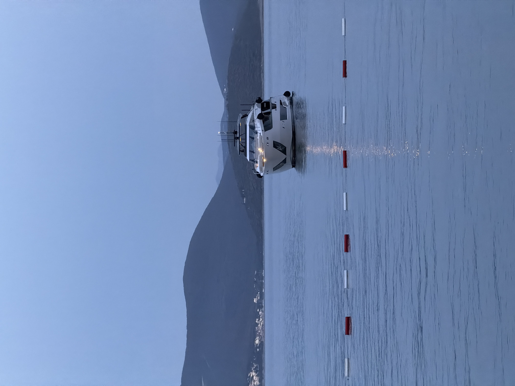 The image depicts a yacht anchored in the waters of Glavaticici, Montenegro. Mountains line the background, while the sky is a muted blue. A series of red and white buoys mark a swimming area or navigational channel alongside the yacht. The water surface is reflective, with subtle ripples. The image was taken in the late evening and early morning.