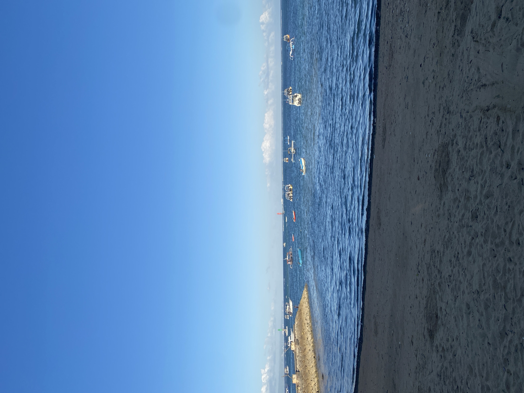 A wide-angle, eye-level, outdoor, full shot captures a serene beach scene in Denpasar, Indonesia, under a clear, bright blue sky. The foreground is dominated by a sandy beach, with subtle textures and indentations indicating the passage of time. To the left, a rocky embankment juts out into the calm, blue ocean, forming a protective barrier.
The middle ground is a expanse of deep blue water, reflecting the sky and dotted with numerous boats of various sizes and types. Some are small, traditional fishing vessels with outriggers, while others appear to be motorboats. A few boats are anchored near the rocky embankment, and others are more scattered across the water. The surface of the water is gently rippled, suggesting a slight breeze.
In the distance, a hazy horizon line separates the sea from the sky, with a few wispy white clouds visible near the horizon. The overall impression is one of tranquility and a typical coastal day in Denpasar. There are no visible people or animals in the frame. The lighting is bright, indicating it is likely daytime, possibly late morning or early afternoon given the intensity of the sunlight.