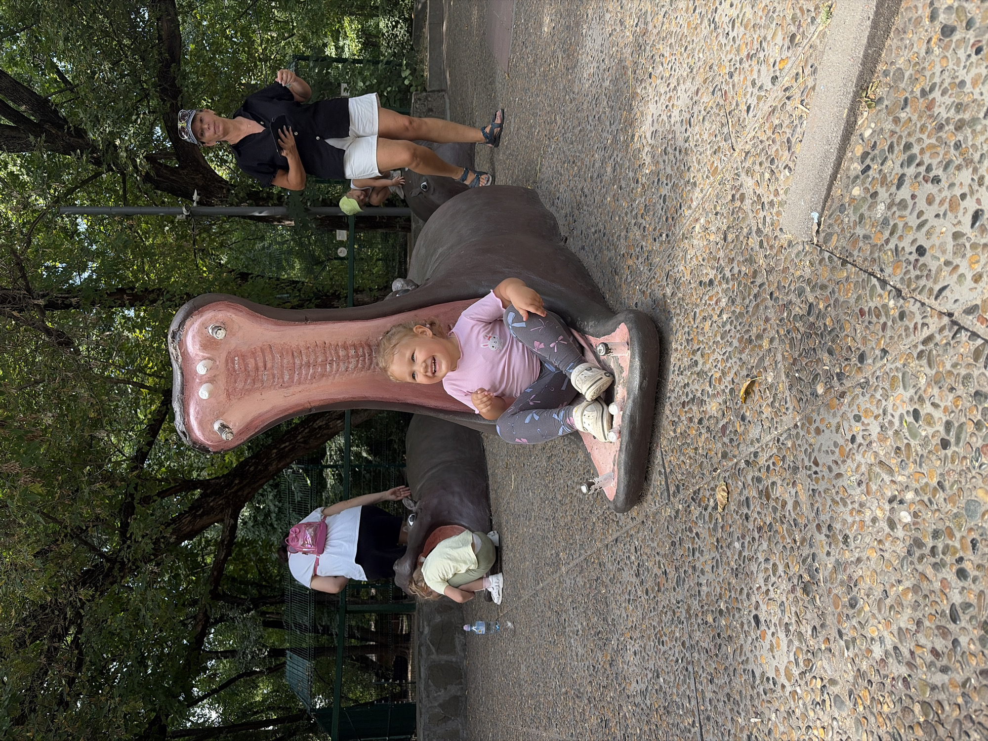 A blonde-haired toddler with a pink shirt and grey leggings is sitting inside the open mouth of a large, grey, hippo-shaped sculpture. She is smiling and looking to her right. The sculpture is located outdoors on a pebble-covered ground. Behind her, another child in a yellow shirt and green pants is crouching down, seemingly interacting with the sculpture. Further back, an adult woman in a black shirt and white shorts stands holding a tablet and a pen, looking towards the camera. A woman in a white shirt and pink backpack is also visible in the background. The environment is a park-like setting with trees providing shade. The scene appears to be at the Rostov-on-Don zoo in Russia, given the presence of a large animal sculpture.