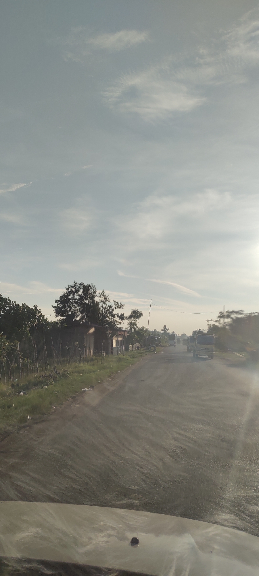 A wide-angle, eye-level shot captures a dusty, rural road under a hazy, pale blue sky with wispy white clouds. The road, which appears to be unpaved or undergoing construction, stretches into the distance, with tire tracks creating a striped pattern on the surface. To the left of the road, a line of lush green vegetation borders a small, dilapidated shack. Beyond the shack, several trees with dense canopies rise, casting shadows. In the middle ground, a few trucks are visible on the road, their forms softened by the atmospheric haze. The sun, positioned on the right side of the frame, casts a bright glare that washes out details in the distance. The foreground shows the hood of a vehicle, suggesting the image was taken from inside a car. This scene is likely located in the city of Meunasahmesjid, Indonesia, given the context.
