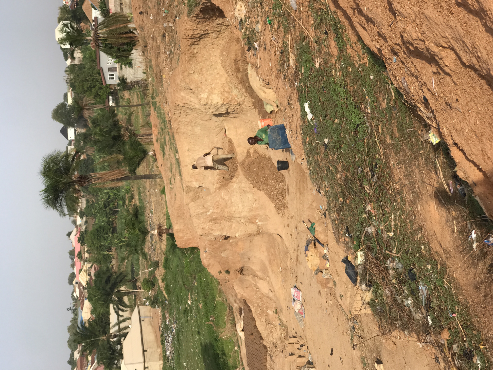 This outdoor image depicts a scene of manual earthwork and residential structures, likely in a peri-urban or developing area, consistent with the context of Abuja, Nigeria. The photograph appears to be taken during daylight hours under a clear sky, with the sun casting distinct shadows.
In the foreground and midground, two individuals are engaged in activity on a large patch of reddish-brown earth. One person, wearing light-colored clothing, is bent over, seemingly digging or shaping the earth with a tool that resembles a shovel or pickaxe. Close by, another person, dressed in a green top, stands next to a large blue plastic container and an orange bucket, appearing to be involved in the collection or processing of the excavated material. The terrain around them is uneven, with dug-out sections forming small, steep banks of earth. Scattered across the ground, particularly along the edges of the dirt path and grassy areas, is a notable amount of litter, including plastic bags, bottles, and other debris.
Beyond the immediate work area, patches of green vegetation and several tall palm trees are visible. In the background, numerous residential buildings with various roof styles and colors, including white, red, and blue, stretch across the landscape, suggesting a populated settlement. Some buildings appear to be under construction or are simple structures. The overall impression is one of human activity shaping the natural environment, possibly for construction purposes or resource extraction. No discernible text is visible in the image.