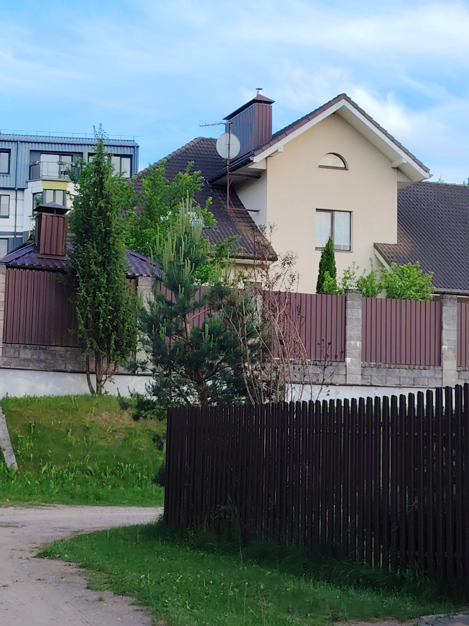 The image presents a quiet residential street scene in Бараўлянскі сельскі Савет, Belarus, under a clear blue sky with sparse white clouds, indicating daytime. The main subject is a two-story house with light beige walls and a dark brown gabled roof. Notable architectural features include a distinctive arched window positioned above a rectangular one on its front facade, a prominent chimney, and a satellite dish mounted on the roof.
To the left and slightly behind, a more modern-looking building with a flat roof, balconies, and grey corrugated siding is partially visible. Another smaller building with a brown roof and chimney is situated behind the left section of the main property's fence.
The properties are enclosed by two fences. In the mid-ground, a tall brown fence made of corrugated metal panels set between grey concrete or stone pillars stretches across the scene. In the foreground, a lower, dark brown wooden picket fence runs along the edge of a dirt or gravel path that curves through patches of green grass.
Various types of vegetation populate the scene. Tall, slender coniferous trees stand alongside the fences, while several pine trees, one displaying noticeable new, lighter green growth, are also present. Bare deciduous branches are visible on the left side. Lush green grass covers the ground, including a gentle grassy slope behind the foreground fence. No people are visible, and no specific activities are taking place, portraying a calm, static view of a suburban or rural landscape. There is no visible text in the image.