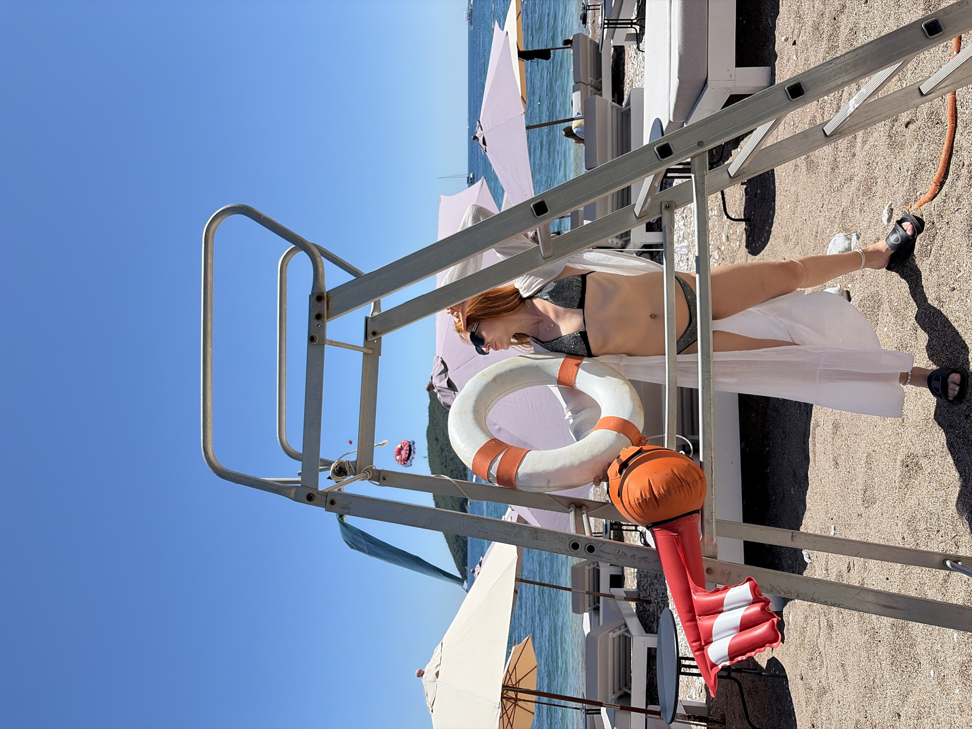 A vibrant beach scene unfolds on a clear, sunny day, featuring a woman relaxing by a lifeguard stand. The bright blue sky fills a significant portion of the image, suggesting midday sun.
In the foreground, the sturdy metal frame of a lifeguard stand, complete with handrails and a ladder, dominates the view. A woman with reddish hair, wearing sunglasses, a dark sequined bikini top, and a flowing white sheer skirt, is seen lying beneath the stand, appearing relaxed. Her bare midriff and legs, ending in black sandals, are visible.
Attached to the lifeguard stand are essential safety items: a white life preserver with orange stripes and a distinctive orange and red striped inflatable marker buoy. The sandy and pebbly beach stretches into the mid-ground, where an orange hose lies.
Beyond the immediate foreground, the sparkling blue sea stretches horizontally, framed by a line of light-colored beach umbrellas and loungers. In the distance, small boats or yachts dot the water, and a paraglider with a red and white canopy floats above a verdant, hilly coastline. The overall impression is one of leisure and summer vacation at a bustling coastal resort, such as those found in Budva, Montenegro. No visible text is present.