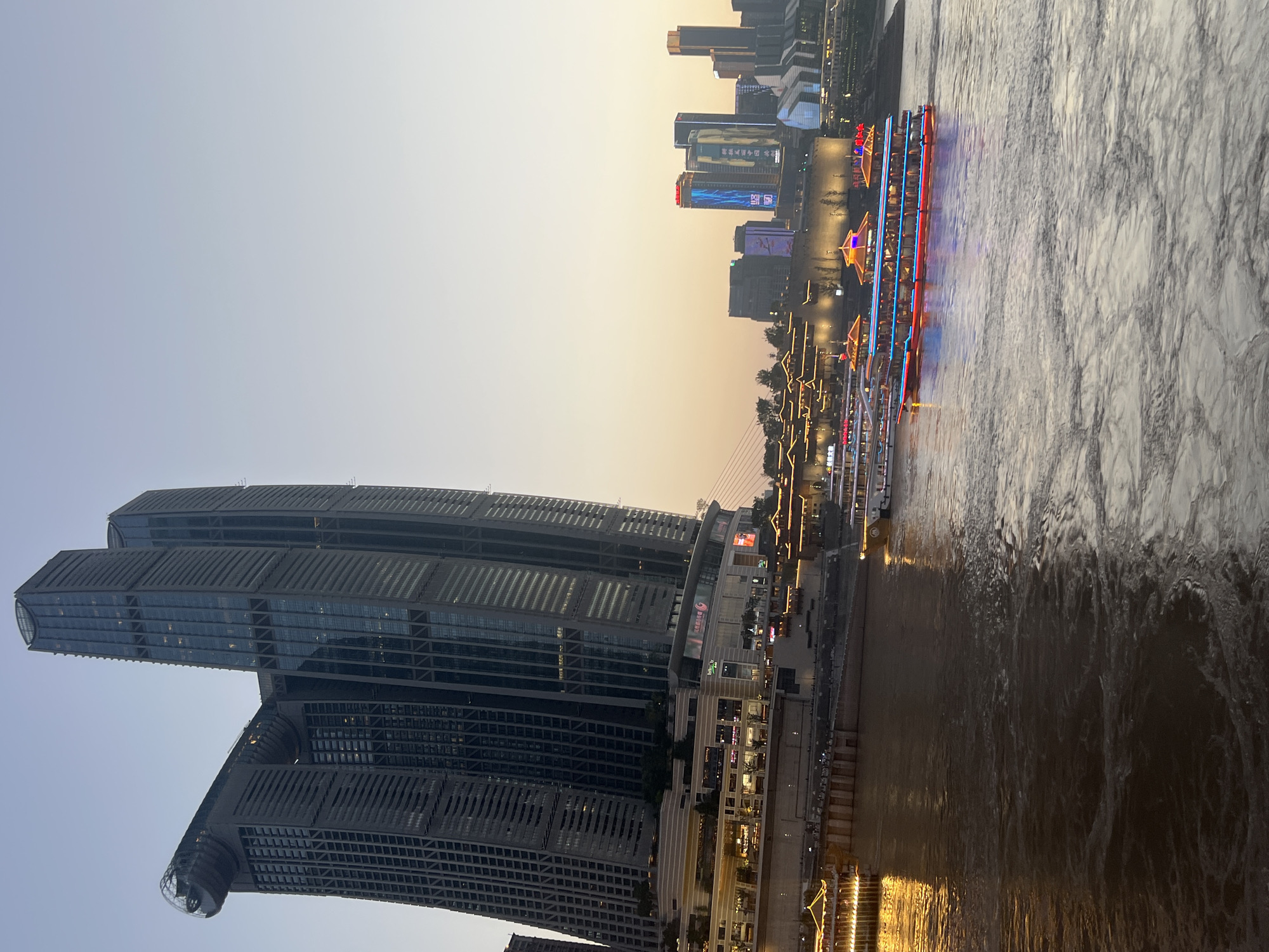 The image depicts a waterfront scene at dusk in Nan'an District, Chongqing, China. Dominating the left side of the frame is the iconic Raffles City Chongqing complex, a multi-tower skyscraper with a distinctive horizontal element connecting two of the towers, resembling a ship's hull. The buildings are illuminated by interior lights and feature a sleek, modern architectural style. Below the skyscrapers, a multi-story shopping mall or commercial complex is visible, with bright lights indicating active businesses.
The foreground is dominated by a wide river, likely the Yangtze River, with significant turbulence and swirling patterns on its surface, suggesting movement from boats or currents. Two passenger boats are navigating the river. The boat closer to the viewer is illuminated with vibrant red, blue, and yellow lights, highlighting its ornate design with multiple decks and pagoda-like structures. Another boat, further in the distance to the left, is also lit, casting a warm golden glow on the water.
Across the river, the cityscape of Chongqing's central business district is visible, characterized by a cluster of illuminated skyscrapers, some featuring large digital billboards displaying glowing Chinese characters and images. A suspension bridge is faintly visible in the background, spanning the river. The sky is a soft gradient of pale blue to light orange, indicating the twilight hour. No individuals are clearly discernible in the image.