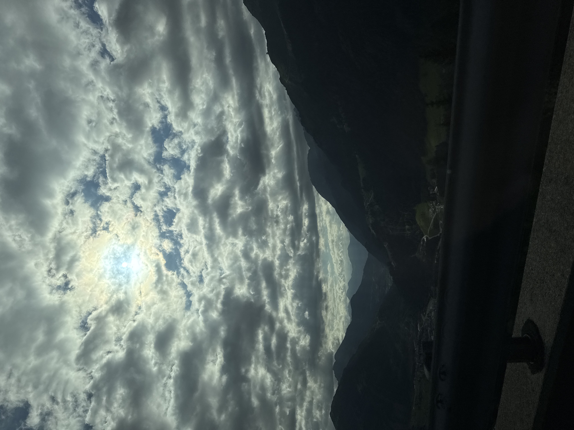The image depicts a scenic view of a mountainous landscape, likely taken from an elevated position such as a road or lookout point, given the presence of a dark railing in the foreground. The sky is filled with dramatic clouds, with the sun peeking through in the upper center, creating a bright, almost halo-like effect.
Below the clouds, a series of dark, silhouetted mountain ranges recede into the distance. The valleys between the mountains appear to be inhabited, with visible signs of human settlement including buildings and possibly roads, suggesting a location near a town or village. The overall atmosphere is moody and somewhat overcast, with the sunlight providing a strong contrast against the darker, shadowed areas of the mountains. The green of the vegetation is visible in patches, especially in the valleys.
While the exact location is not explicitly identified in the image itself, the mention of Airolo, Switzerland, would suggest this is a view within or near that region. Airolo is known for its mountainous terrain and is situated in the canton of Ticino, a region that features similar dramatic landscapes. The visual elements in the image are consistent with the characteristics of the Swiss Alps. No people or specific actions are discernible in this landscape photograph.