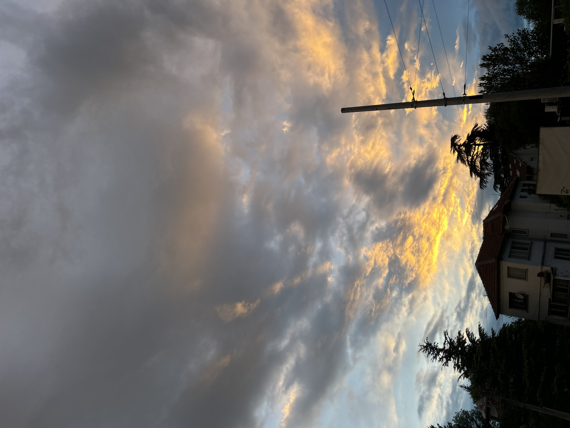 The image shows a late afternoon or early evening sky with layered clouds. Some clouds are dark gray, while others are illuminated with orange and yellow light. The sky transitions from gray to blue. A utility pole with wires stretches across the sky. The silhouette of treetops and a portion of a building with a red-tiled roof are visible at the bottom and right of the frame. The building appears to be an apartment building. The presence of European-style architecture and utility infrastructure suggests the location is most likely in Europe, potentially matching the city of Sofia, Bulgaria. No people, actions, or interactions are explicitly visible.