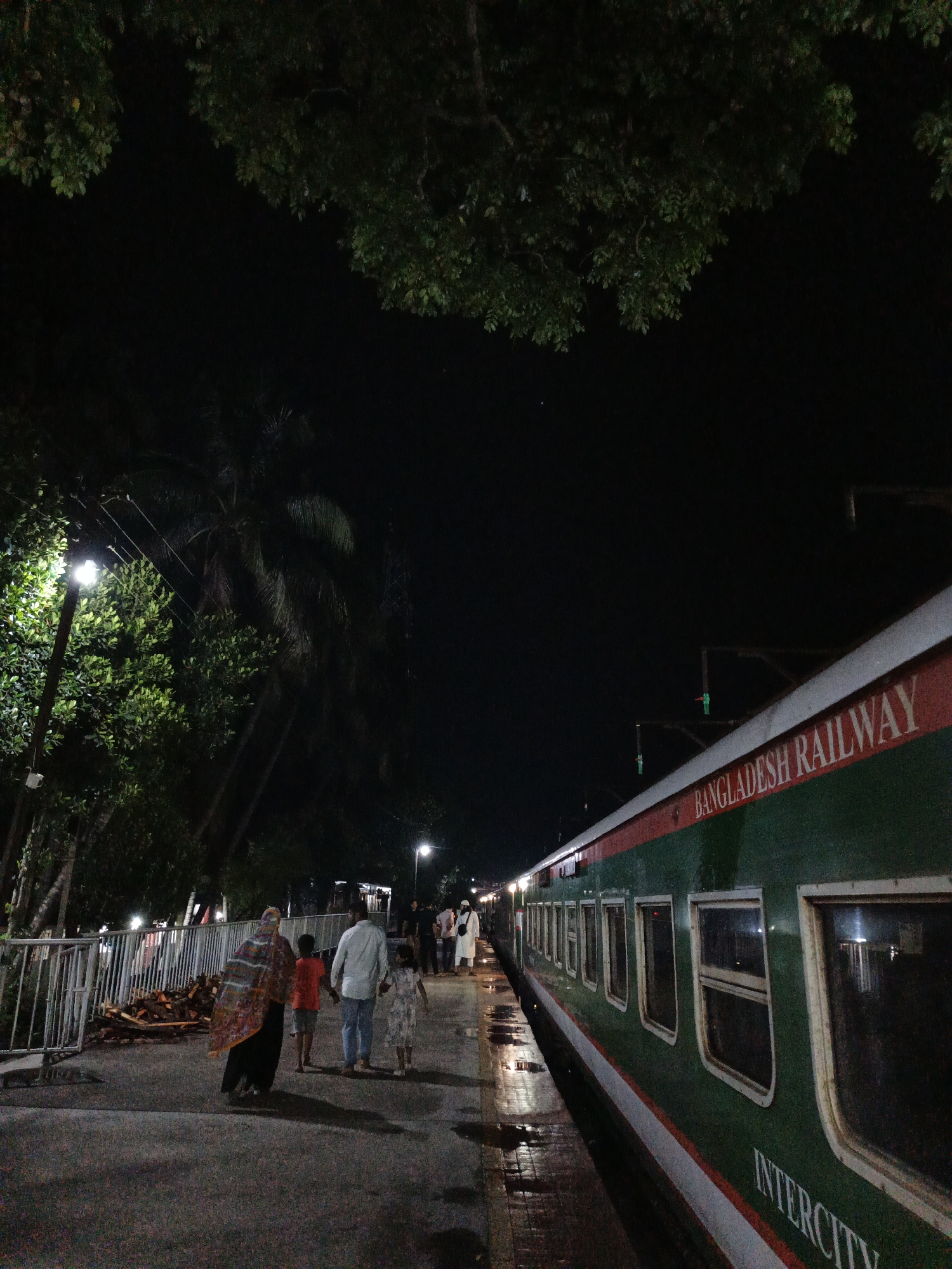 The image depicts a night scene at the Rajbari Railway Station in Rajbari, Bangladesh. A green and red Bangladesh Railway train is parked at the platform. Several people are visible on the platform. In the foreground, a family consisting of two adults and two children are walking away from the camera. The adult on the left, possibly a woman, is wearing a colorful patterned headscarf and a dark outfit. Beside her, a child in an orange shirt walks, followed by an adult male in a white shirt and blue jeans, holding the hand of a child in a light-colored dress. To the right of the train, the platform is wet, reflecting the lights. In the background, more people are seen walking on the platform, and some lights illuminate the area. Trees and foliage are visible on the left side of the image, with a street light illuminating the trees and a section of the platform. The sky is dark, indicating it is nighttime.